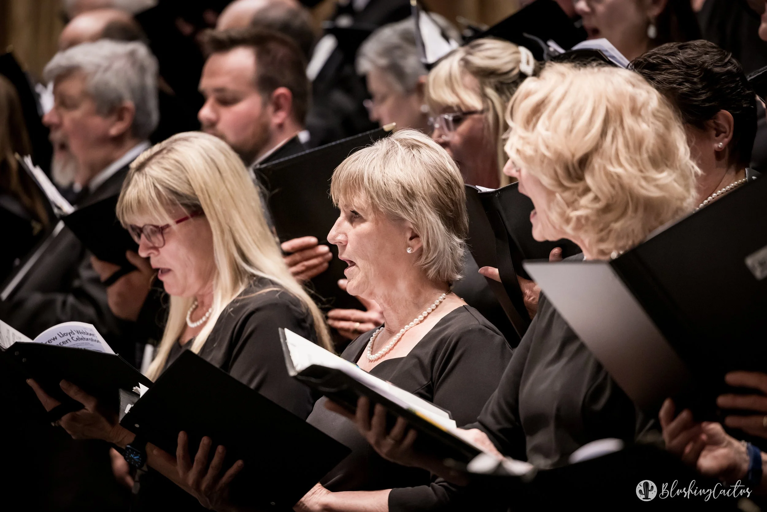 Chorus of people singing in a choir, holding black music folders, with a focus on three women in the front row, all dressed in black and wearing pearl necklaces.