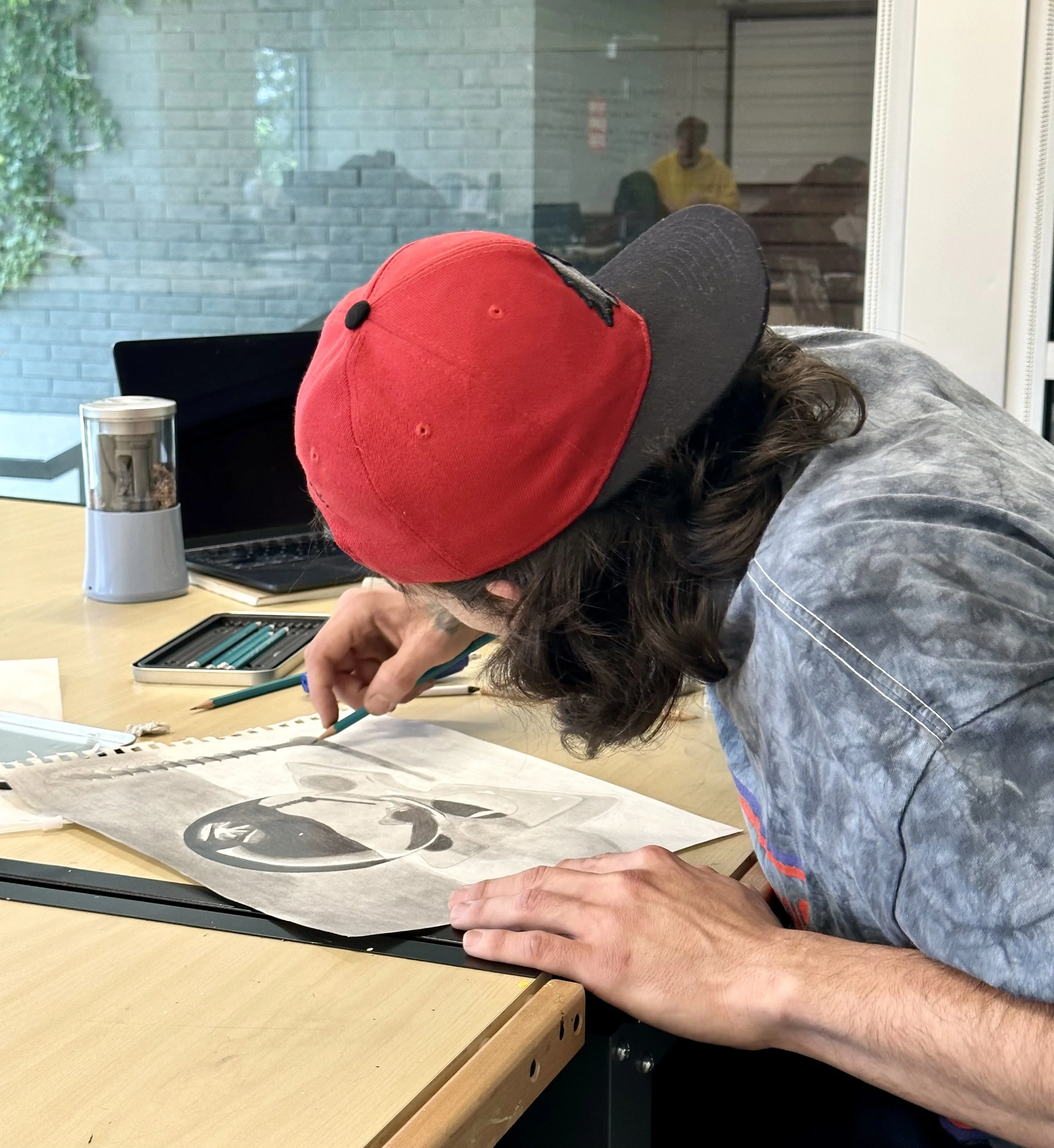 Person with long dark hair wearing a red and black baseball cap and a tie-dye shirt, drawing or working on an art project at a table.