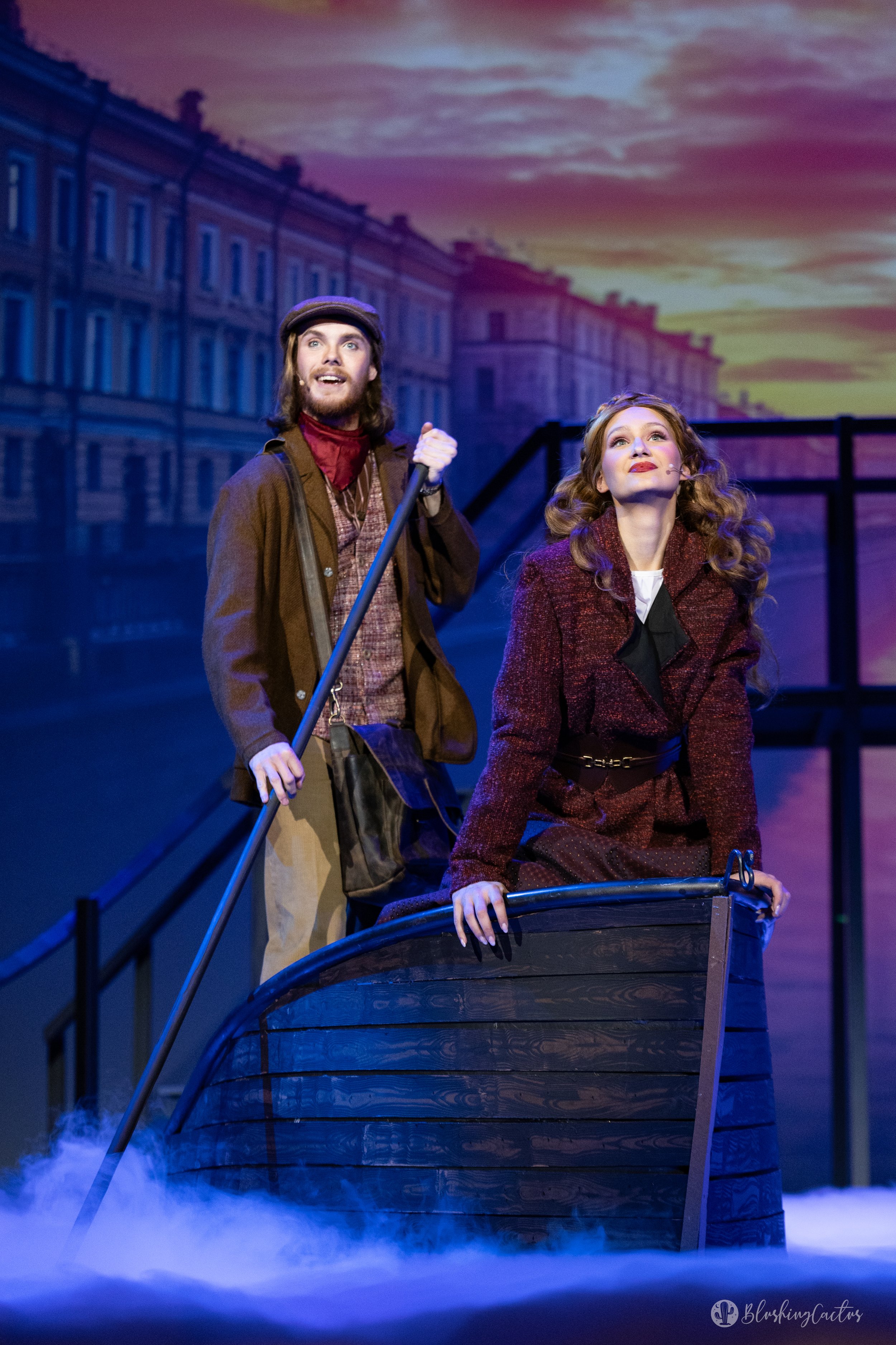 Two actors performing on stage with a backdrop of a cityscape and sky, one man holding a pole and a woman leaning on a boat, suggesting a theatrical scene.