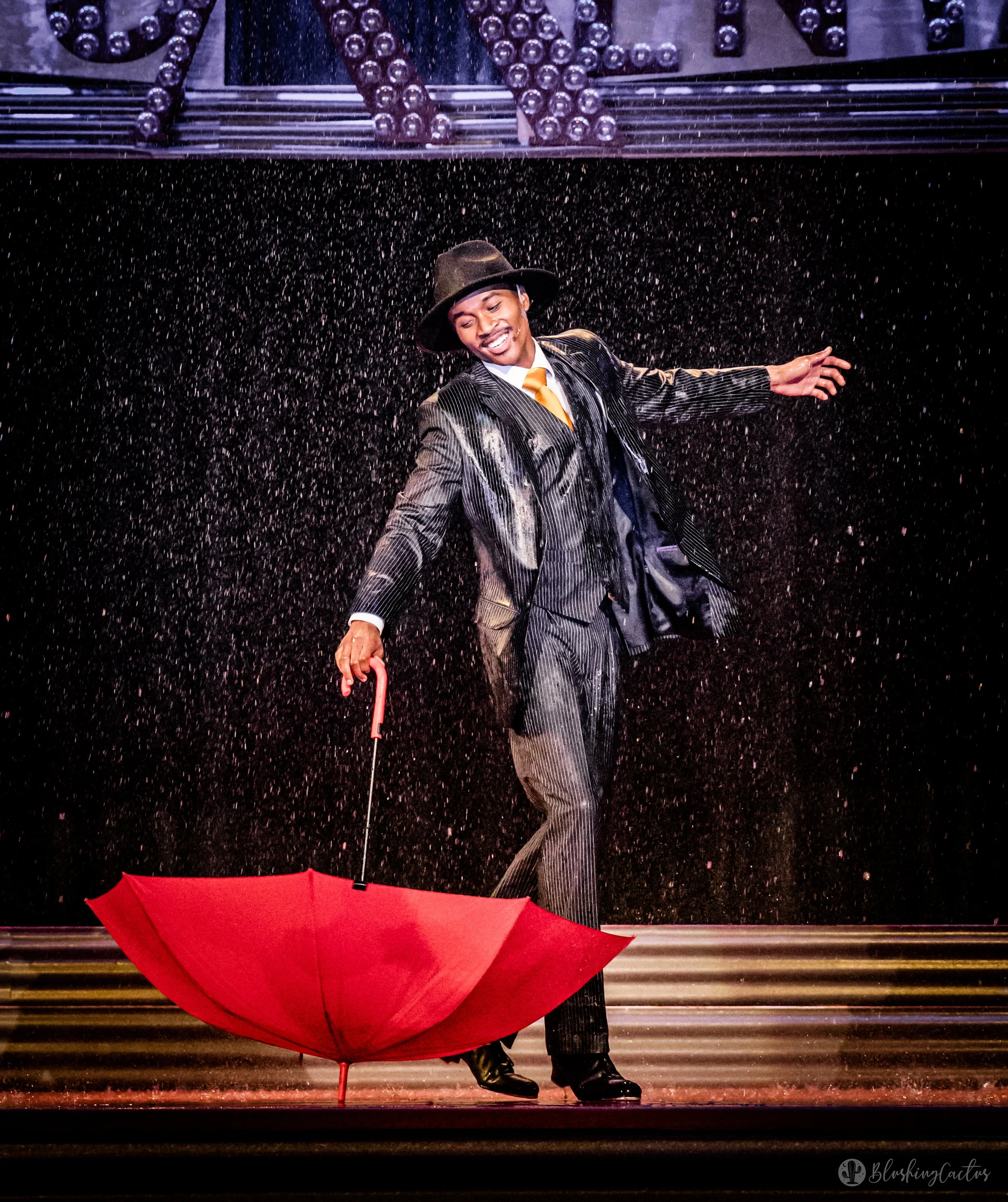 Performer in a suit and hat dancing with an open red umbrella on stage with water spray.