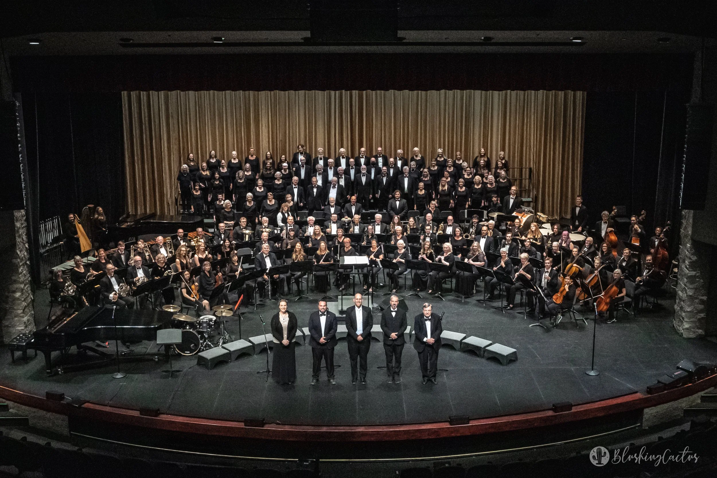 An orchestra on stage with a choir in the background, facing the audience, in a concert hall with a gold curtain and stone pillars, and five people standing in front of the orchestra.