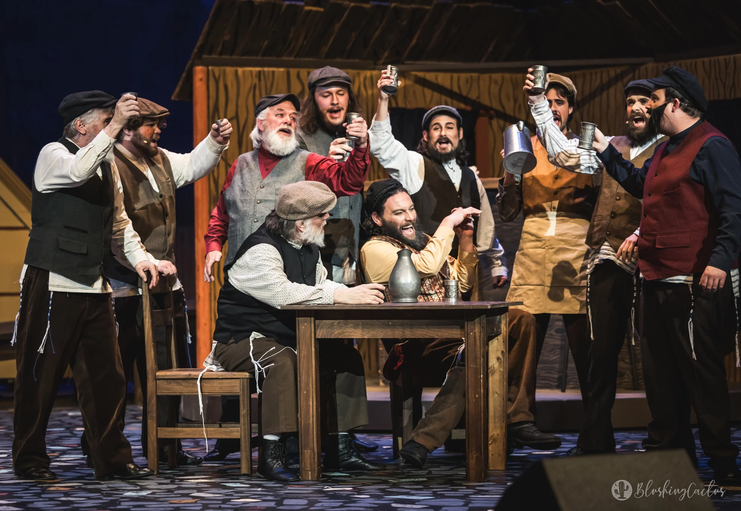 Actors on stage dressed in period costumes, holding up cups in a toast scene from a theatrical play, with a wooden structure in the background.