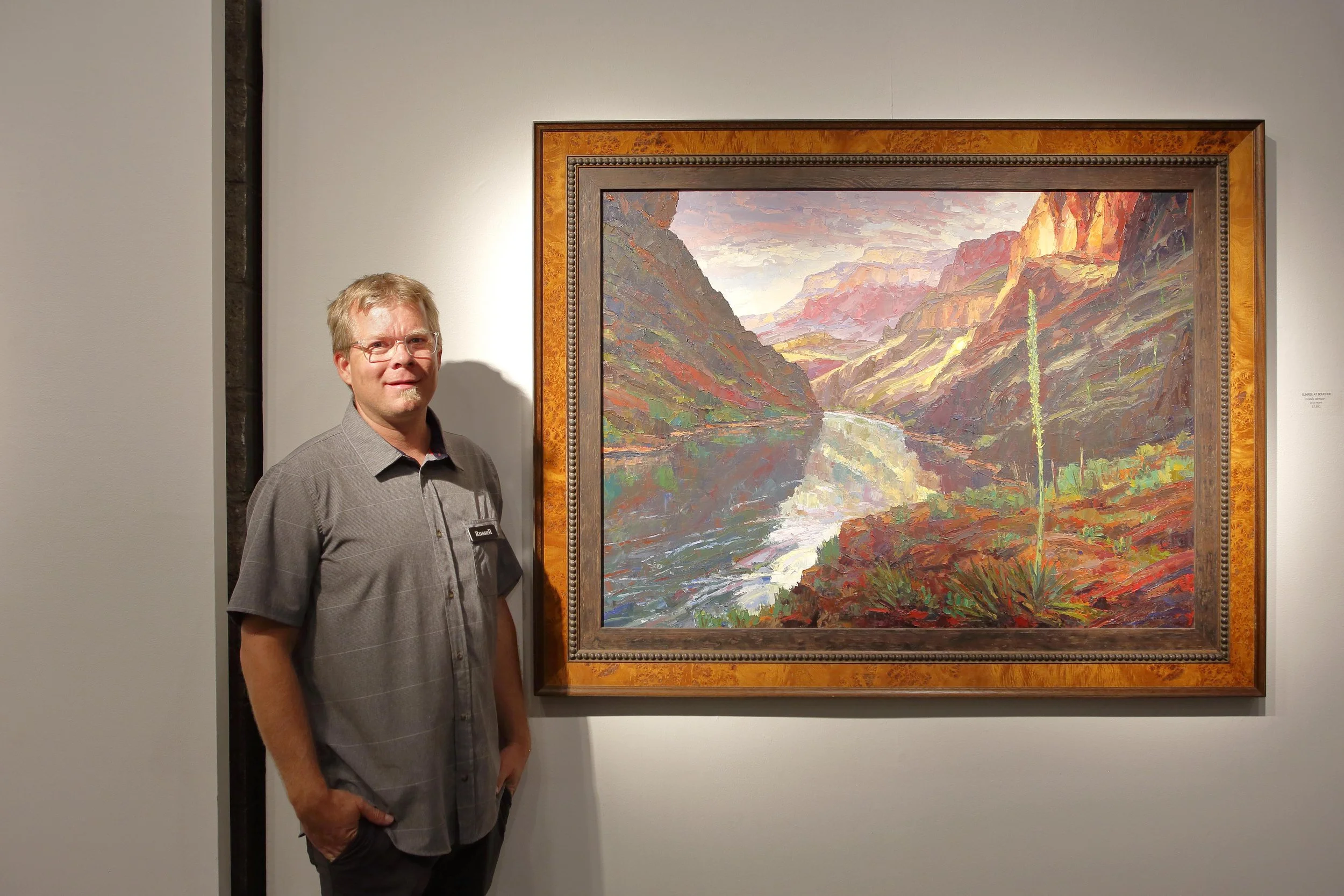 Man standing next to a colorful landscape painting of a canyon with a river, painted in an impressionistic style, displayed in an art gallery.