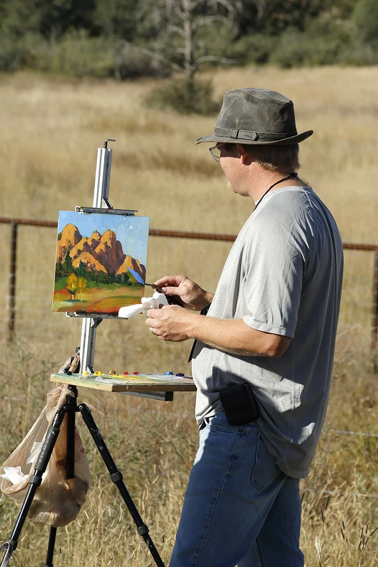 A man wearing a wide-brimmed hat and glasses painting a landscape of mountains and trees on a canvas outdoors, with painting supplies on a portable easel.