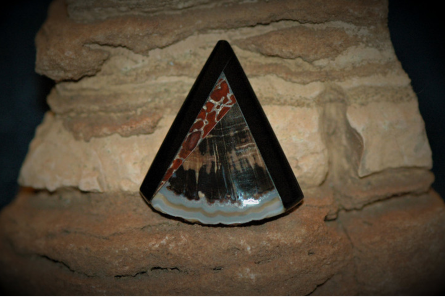 A pyramid-shaped art piece with black, brown, and gray stone patterns, placed on a rough brick surface with a brick wall background.