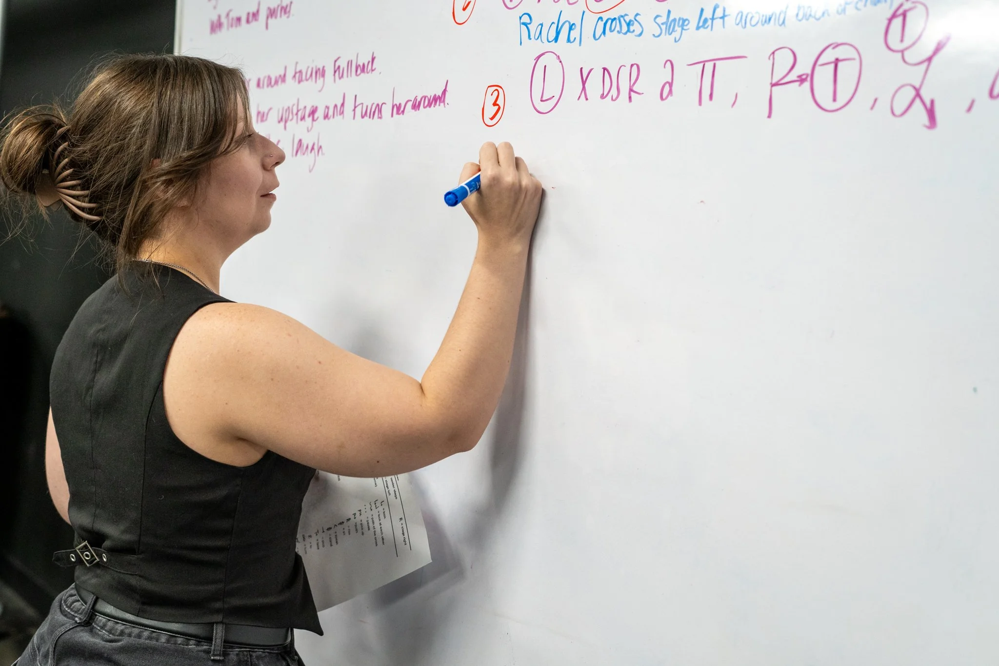 A woman with light brown hair tied in a bun and wearing a black sleeveless top is writing on a whiteboard with a blue marker. The whiteboard has colorful handwritten notes and diagrams, including text in pink, purple, blue, and orange.