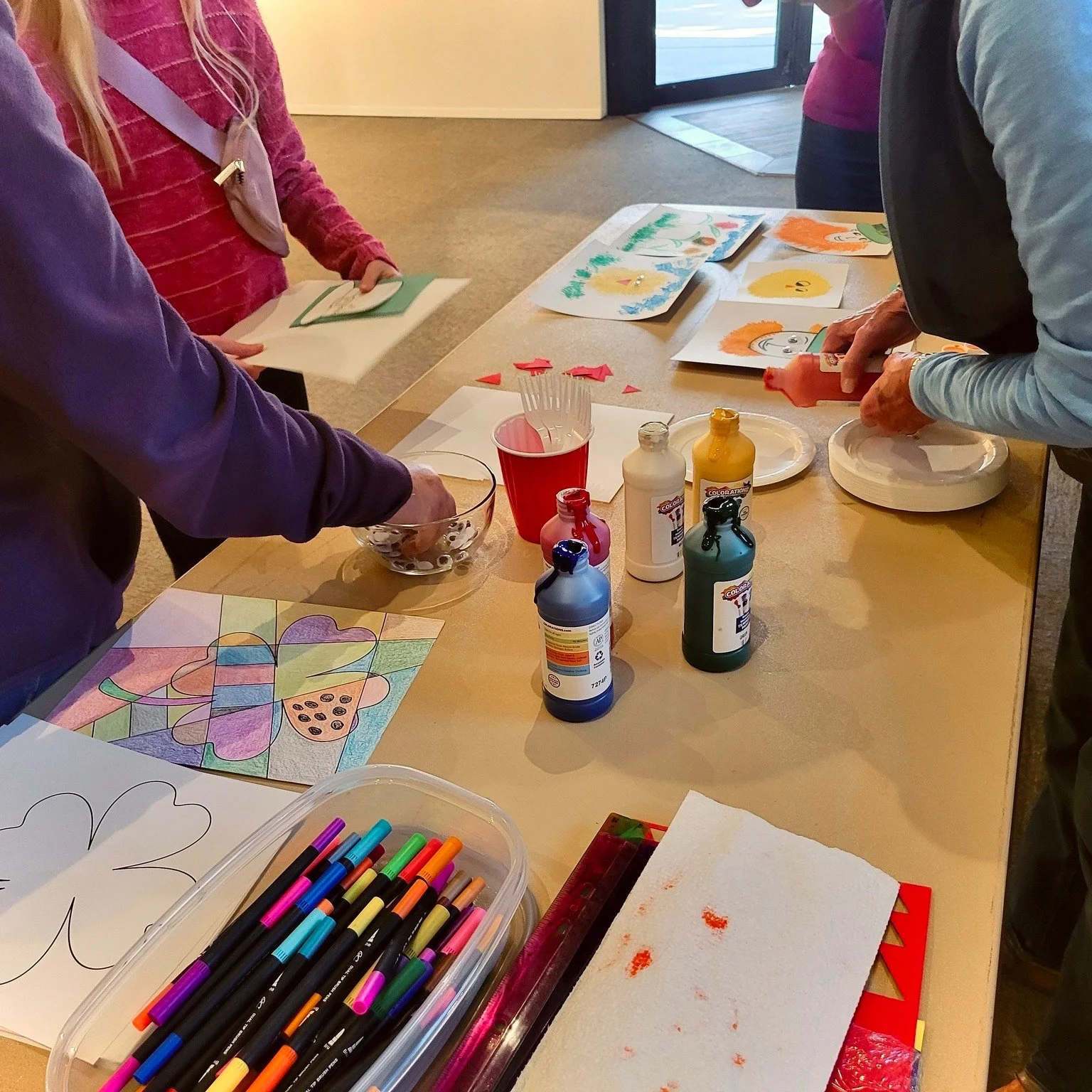 Children and an adult participating in a coloring and craft activity at a table with markers, paints, paper, and artwork.
