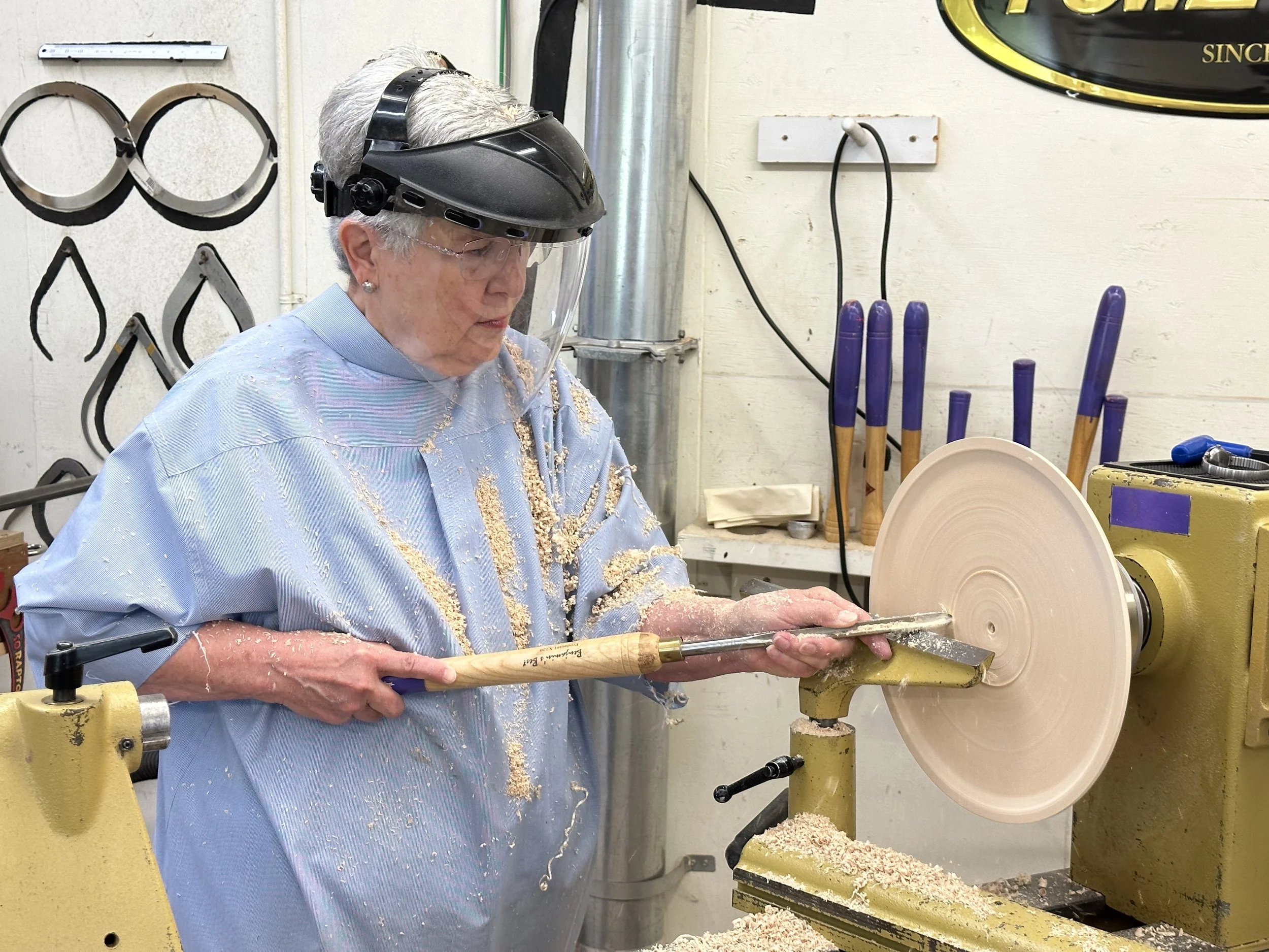 An elderly woman is using a woodturning lathe with a spinning piece of wood, wearing safety equipment including a face shield and safety glasses, in a workshop with wood tools and shavings around.