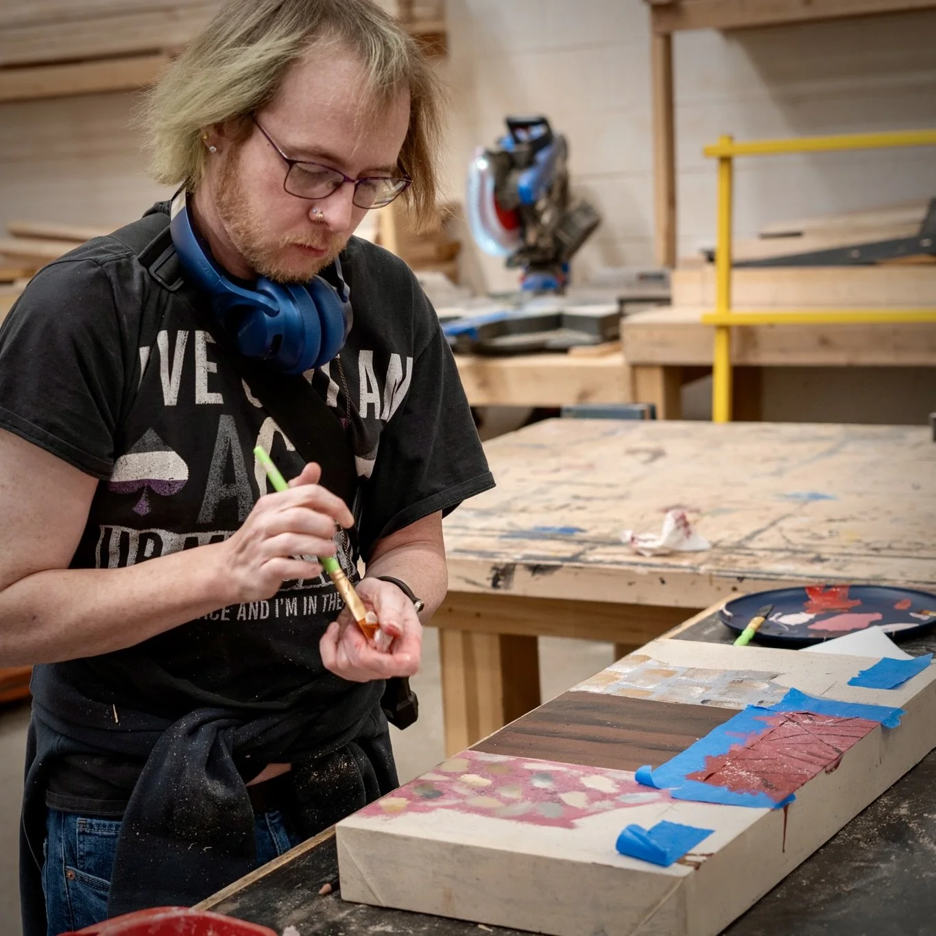 A person with glasses and blue headphones around neck painting a rectangular wooden piece with red paint in a woodworking workshop.