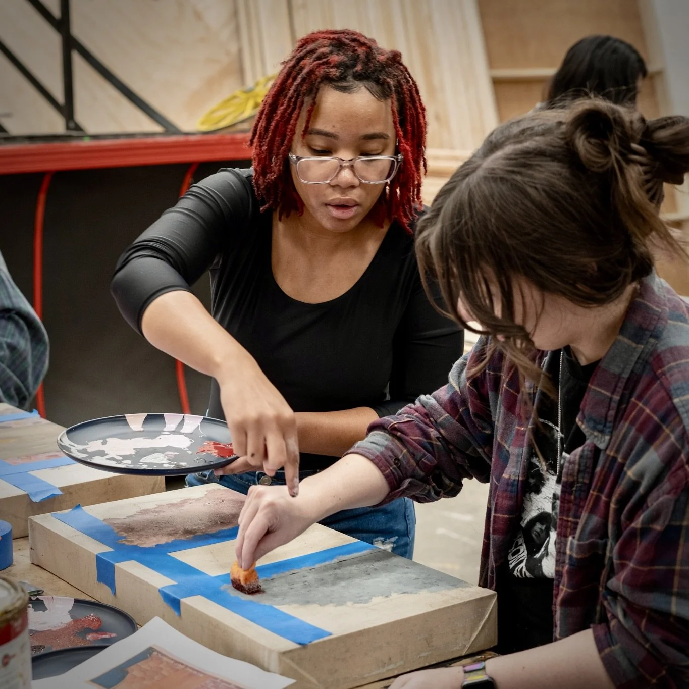 Two women working on an art project, one applying paint with a sponge and the other holding a paint palette, in a workshop setting.