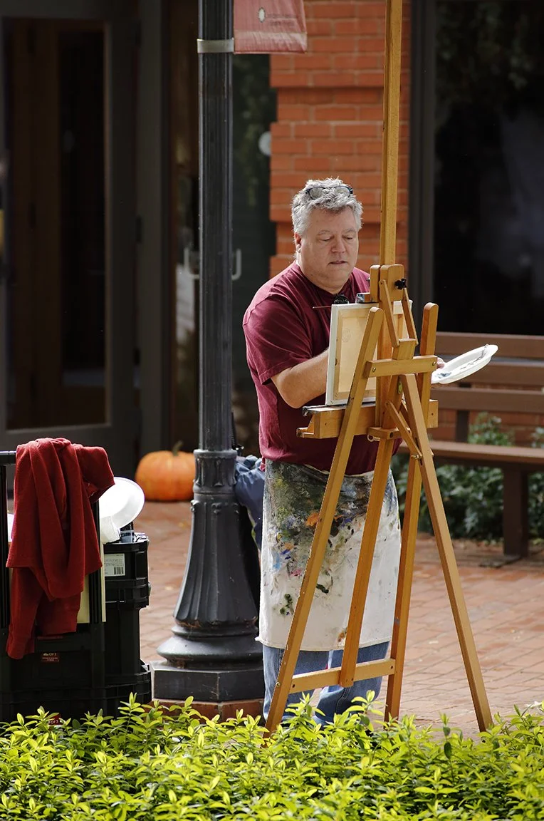 An artist with gray hair, wearing a red shirt and a paint-splattered apron, standing outdoors on a brick patio, painting on a canvas set on an easel, with pumpkins and greenery nearby.