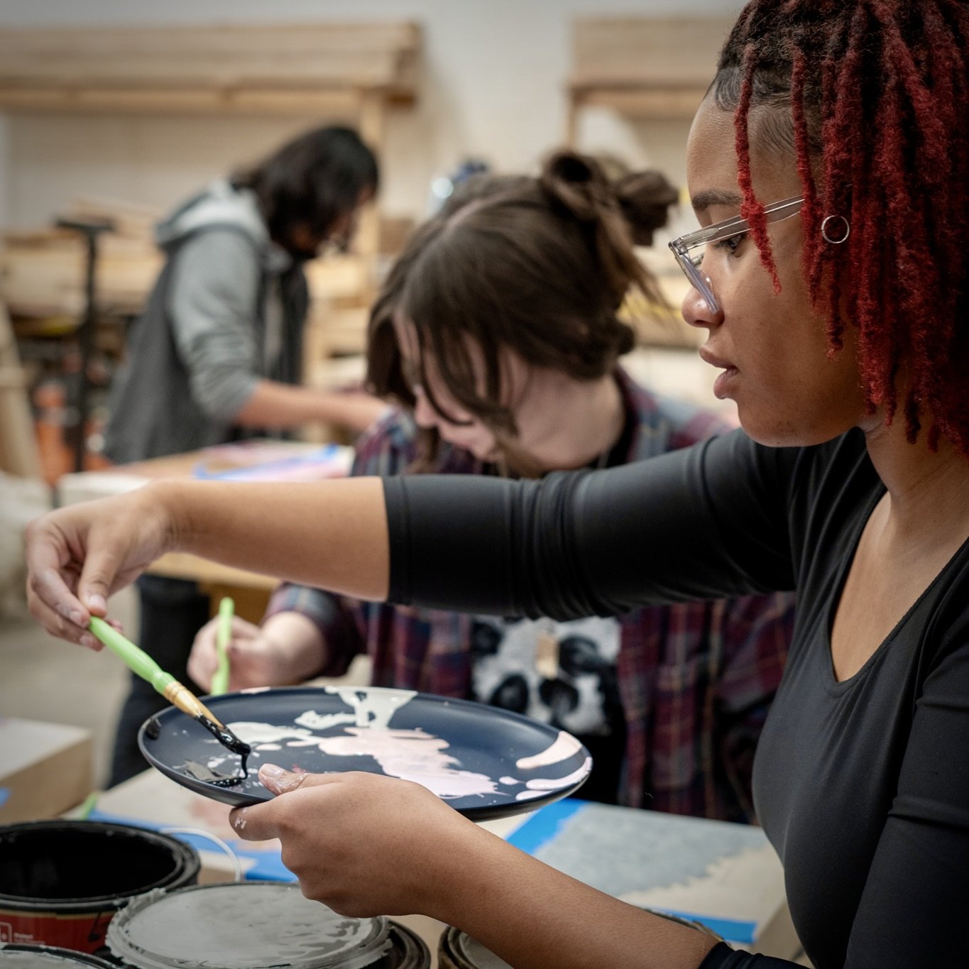 A woman with red dreadlocks and glasses painting with a paintbrush at a workshop, with other people in the background working on their projects.