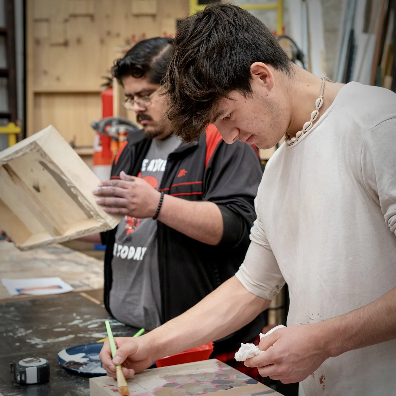 Two young men working on art projects in a woodworking or art studio. One is painting or drawing on a piece of wood, while the other is holding a paper towel.