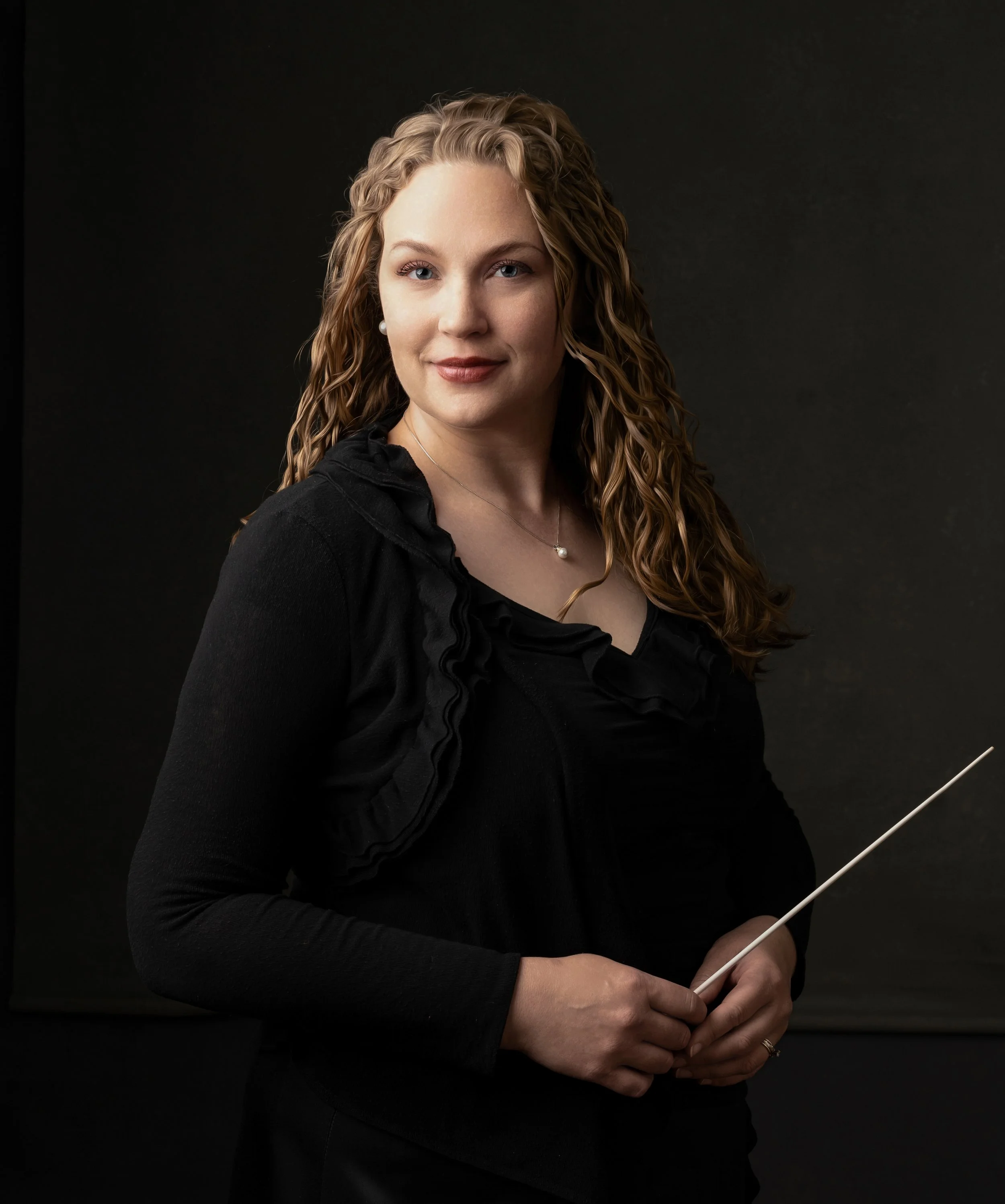 A woman with curly blonde hair, wearing a black top with ruffled details, holding a conductor's baton, standing against a dark background.