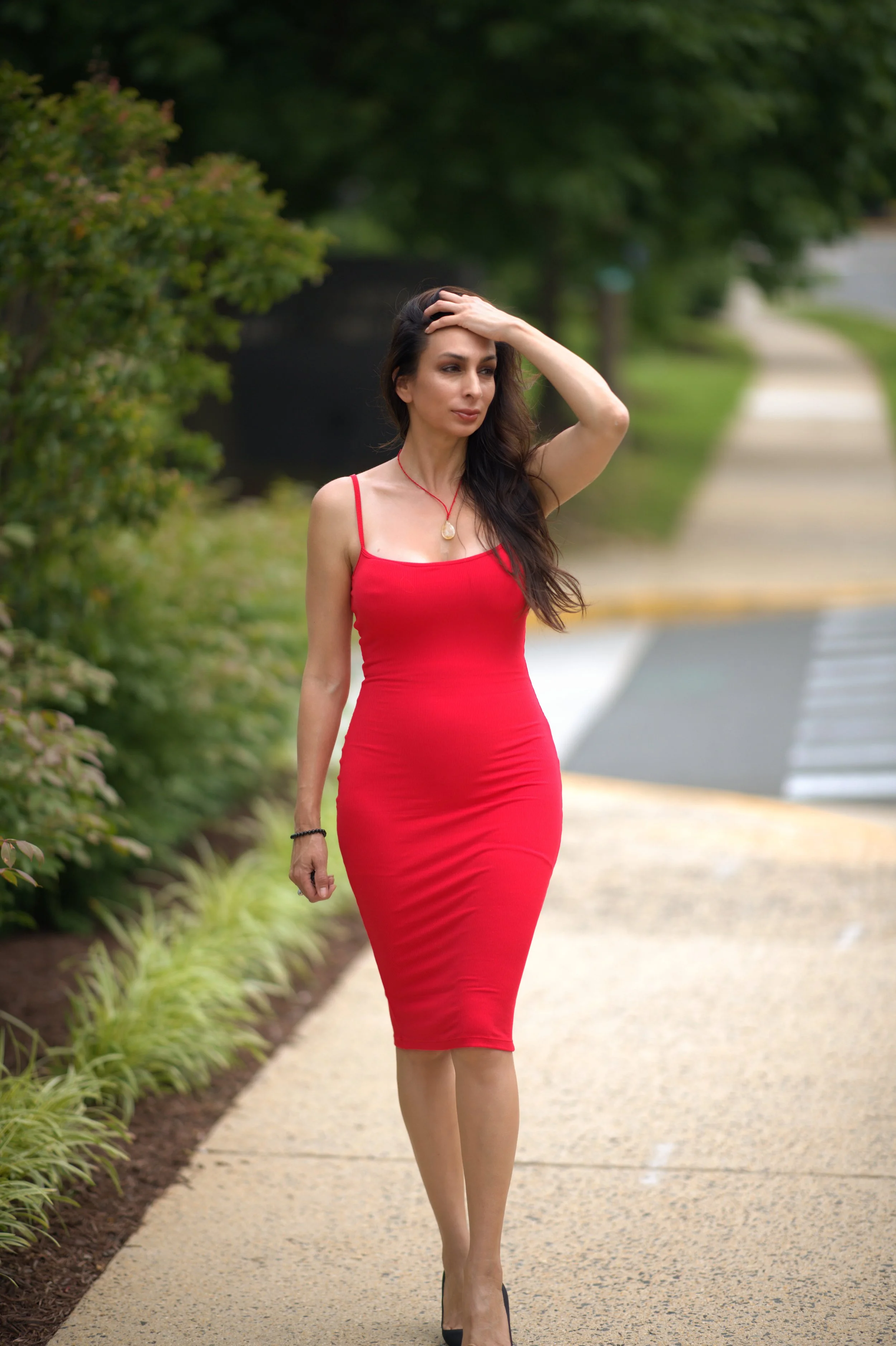 A woman in a red dress walking on a sidewalk with greenery on her side, during daytime.