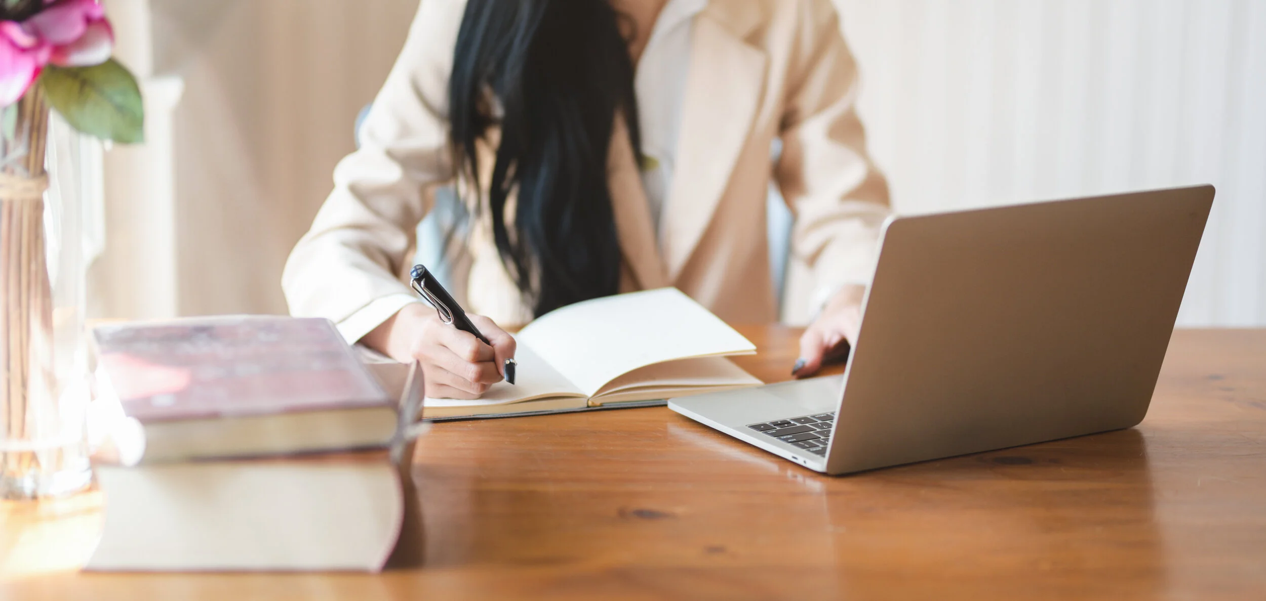 A person sitting at a wooden desk, writing in a notebook with a pen, with a laptop and a stack of books on the side.