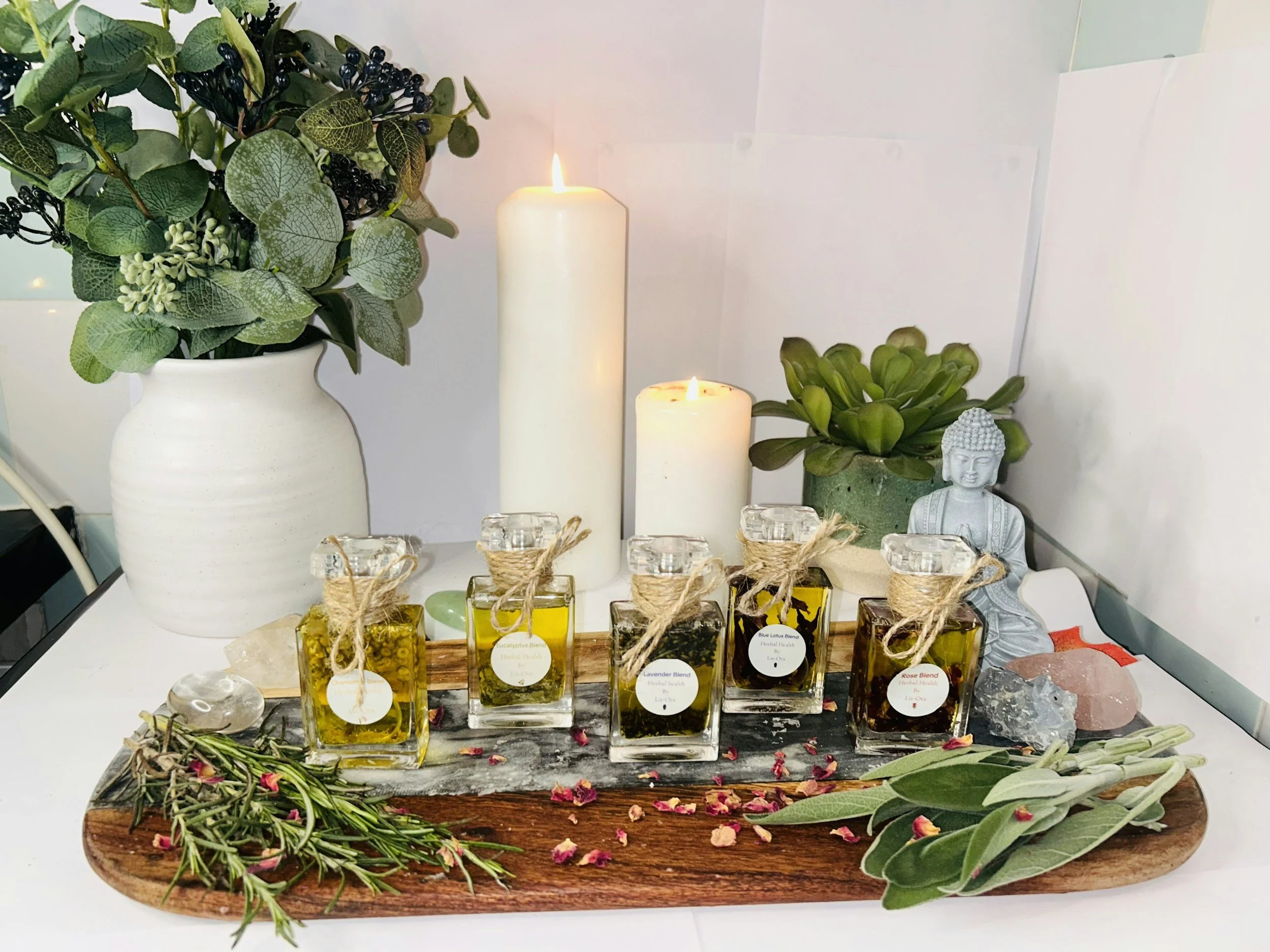 A decorative arrangement with candles, essential oils, greenery, and a Buddha statue on a wooden tray.