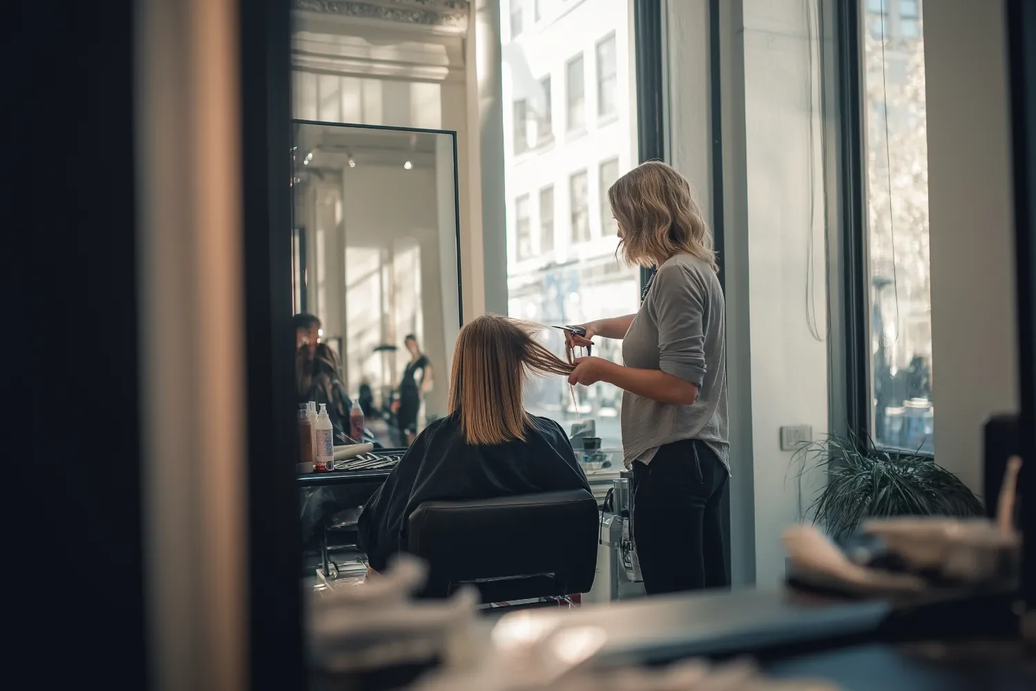 A hairstylist is cutting a woman's hair in a salon with large windows and city view.