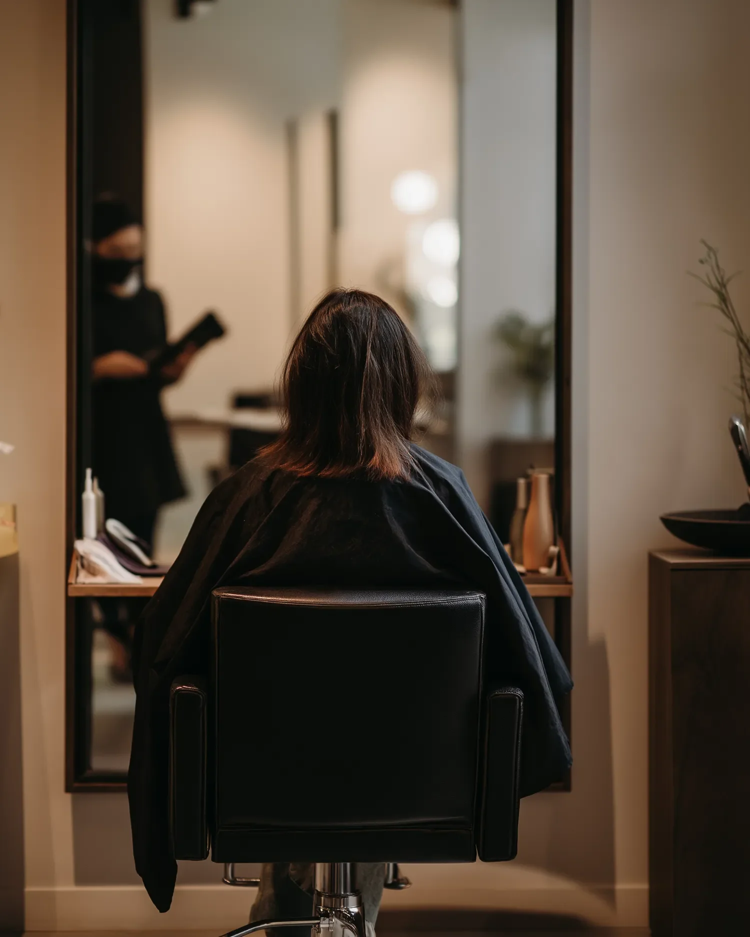 Person sitting in a salon chair at a mirror, preparing for a haircut or styling session. The mirror reflects a stylist working in the background.