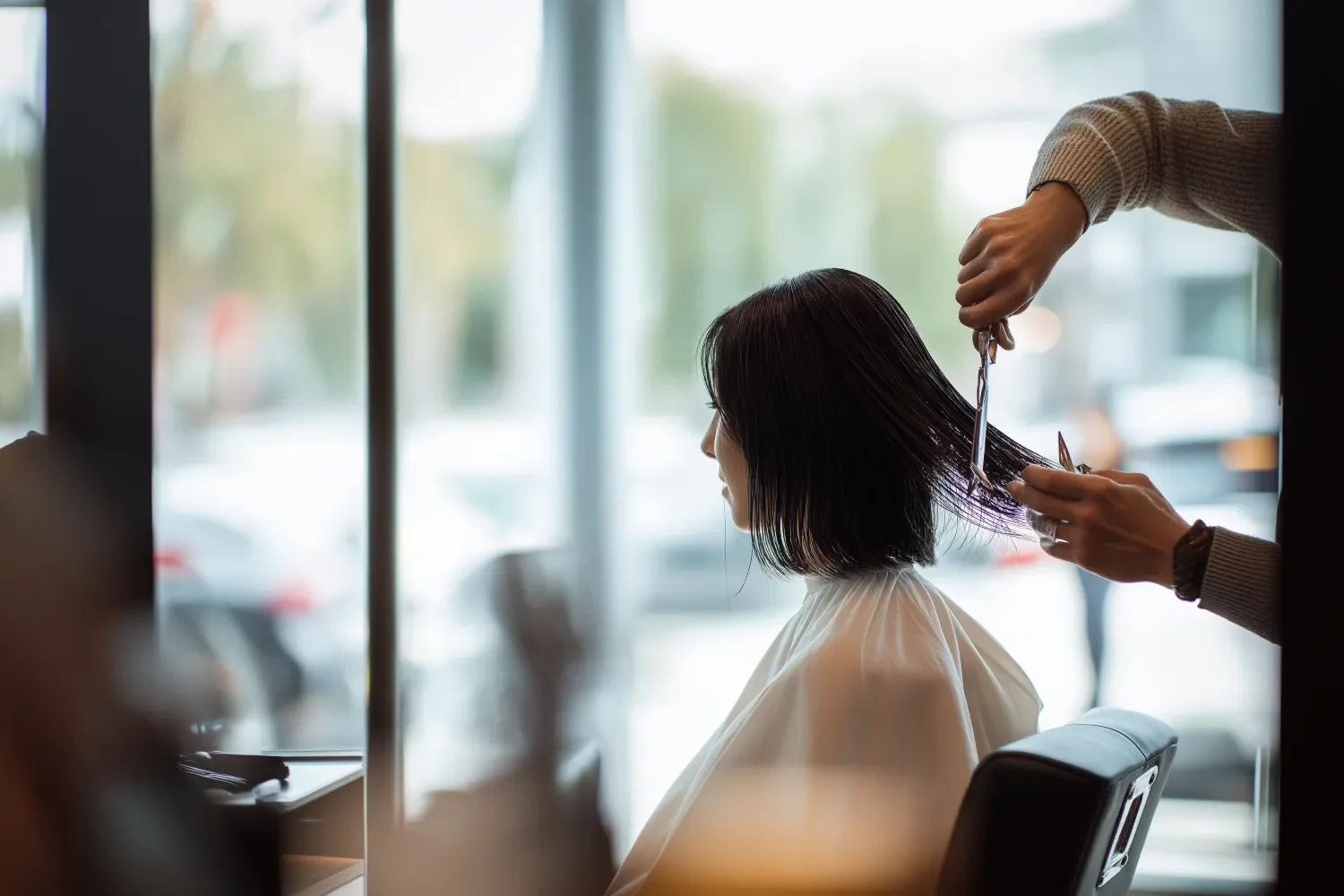 A woman with shoulder-length dark hair getting a haircut at a salon, seated in a salon chair with a cape, while a hairdresser cuts her hair with scissors inside a well-lit salon with large windows.