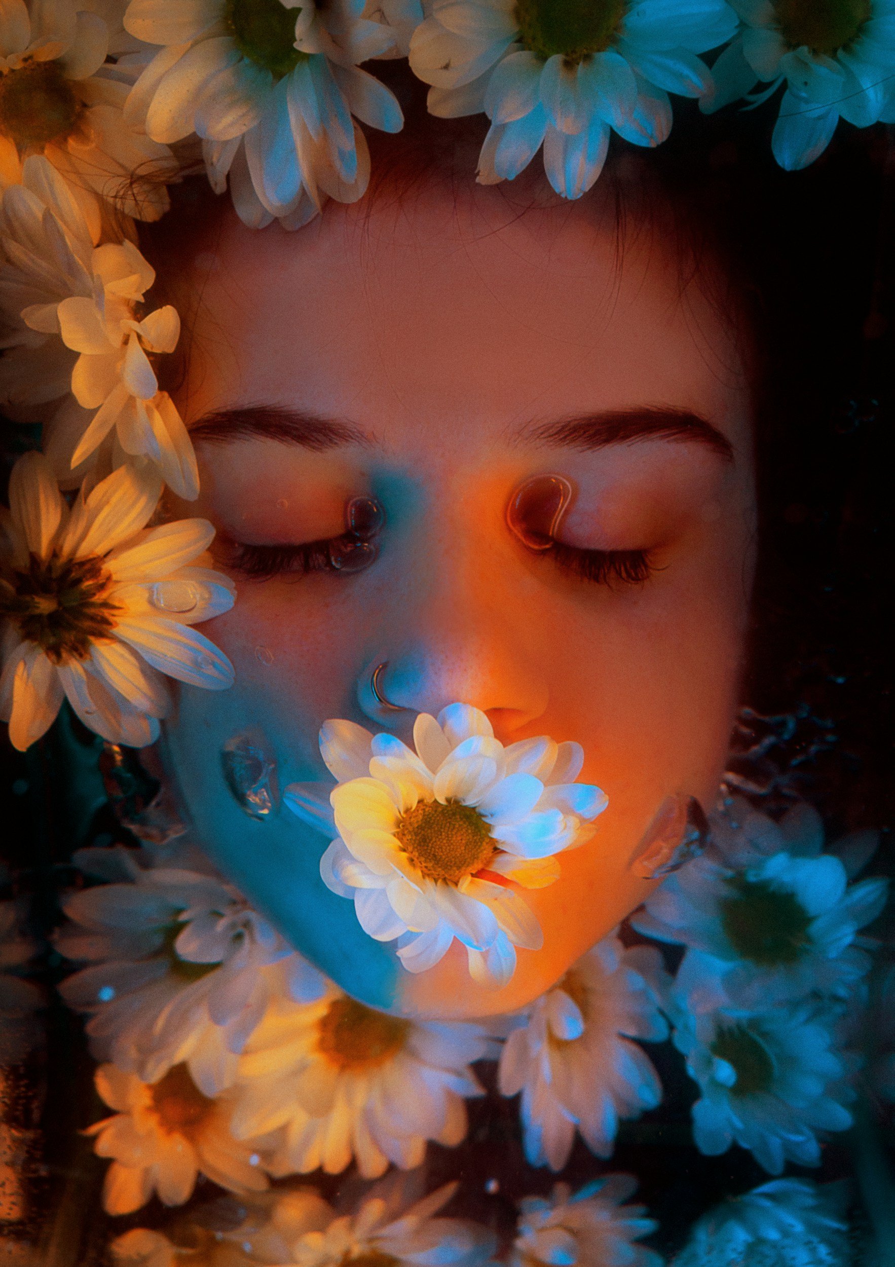 A person with their eyes closed is submerged in water surrounded by white daisies. They have two nose rings and a daisy in their mouth, with bubbles rising from their nose, and a soft, warm light illuminating their face.