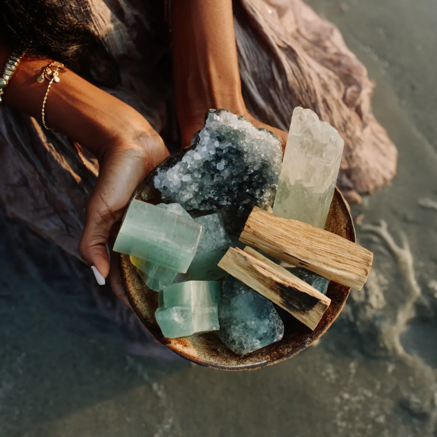 Hand holding a bowl with various large crystals and stones, including gypsum, quartz, and wood pieces, near a beach.