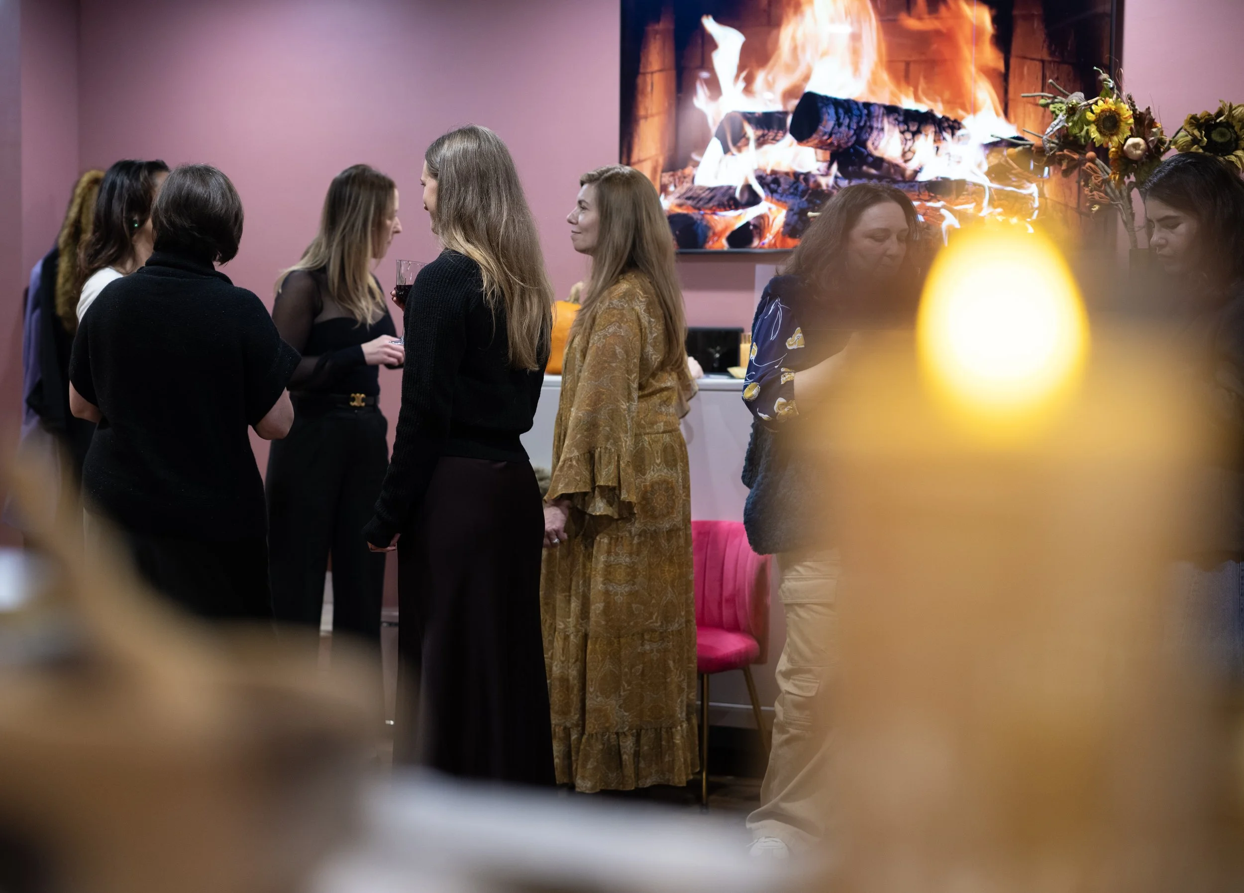 Group of women at a social gathering, standing and chatting in a room with pink walls and a large television screen displaying a fire.