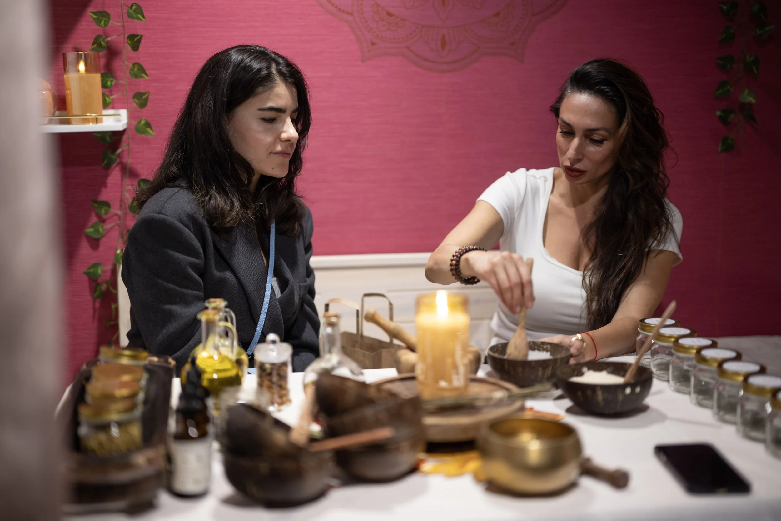 Velta and another woman seated at a table with various herb jars, bowls, and bottles, engaging in a custom made herb based remedy for self care therapy in a room with pink walls and decorative elements.