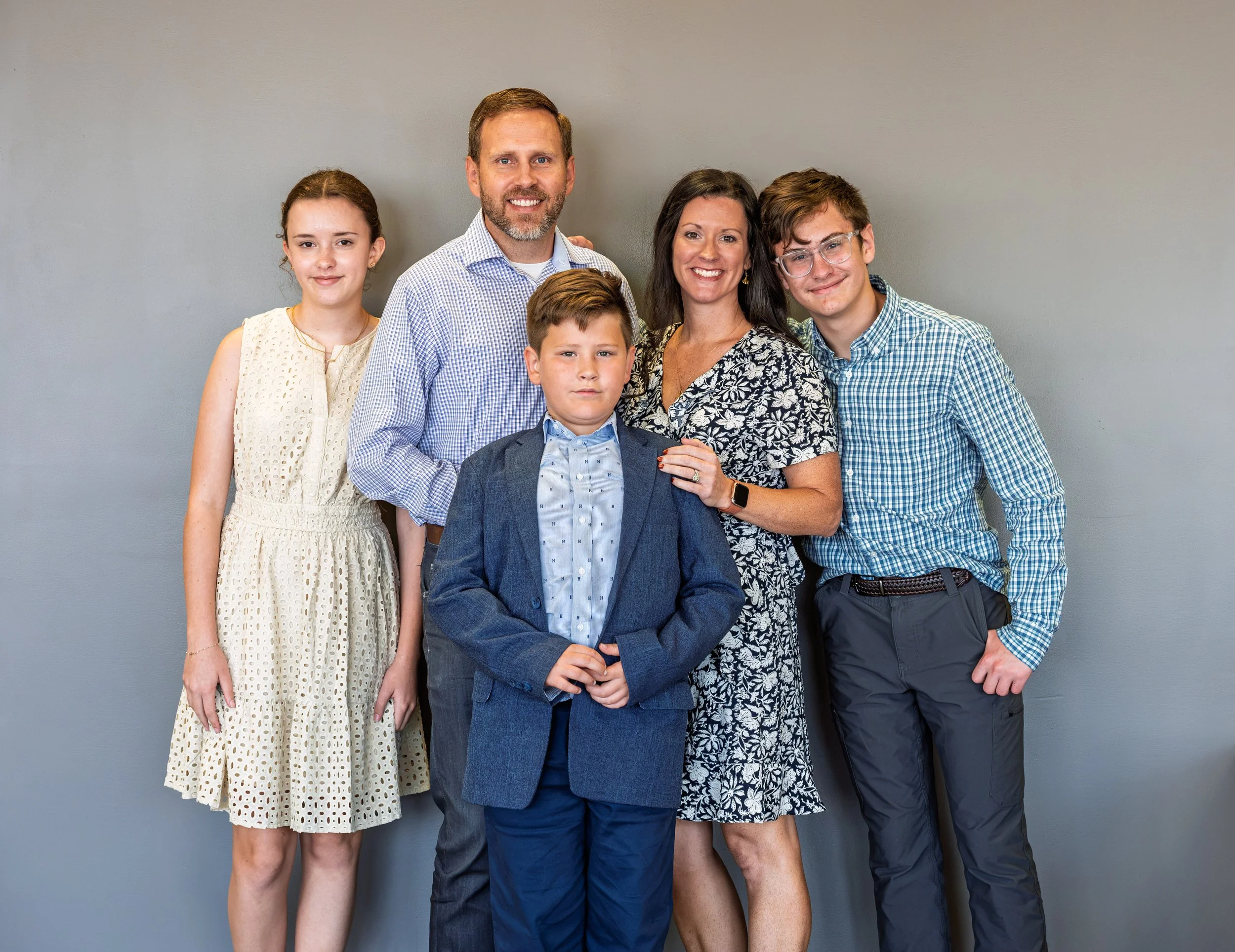 A family of six standing together against a plain gray wall. The group includes two girls, two boys, a man and a woman, all smiling and dressed in semi-formal clothing.