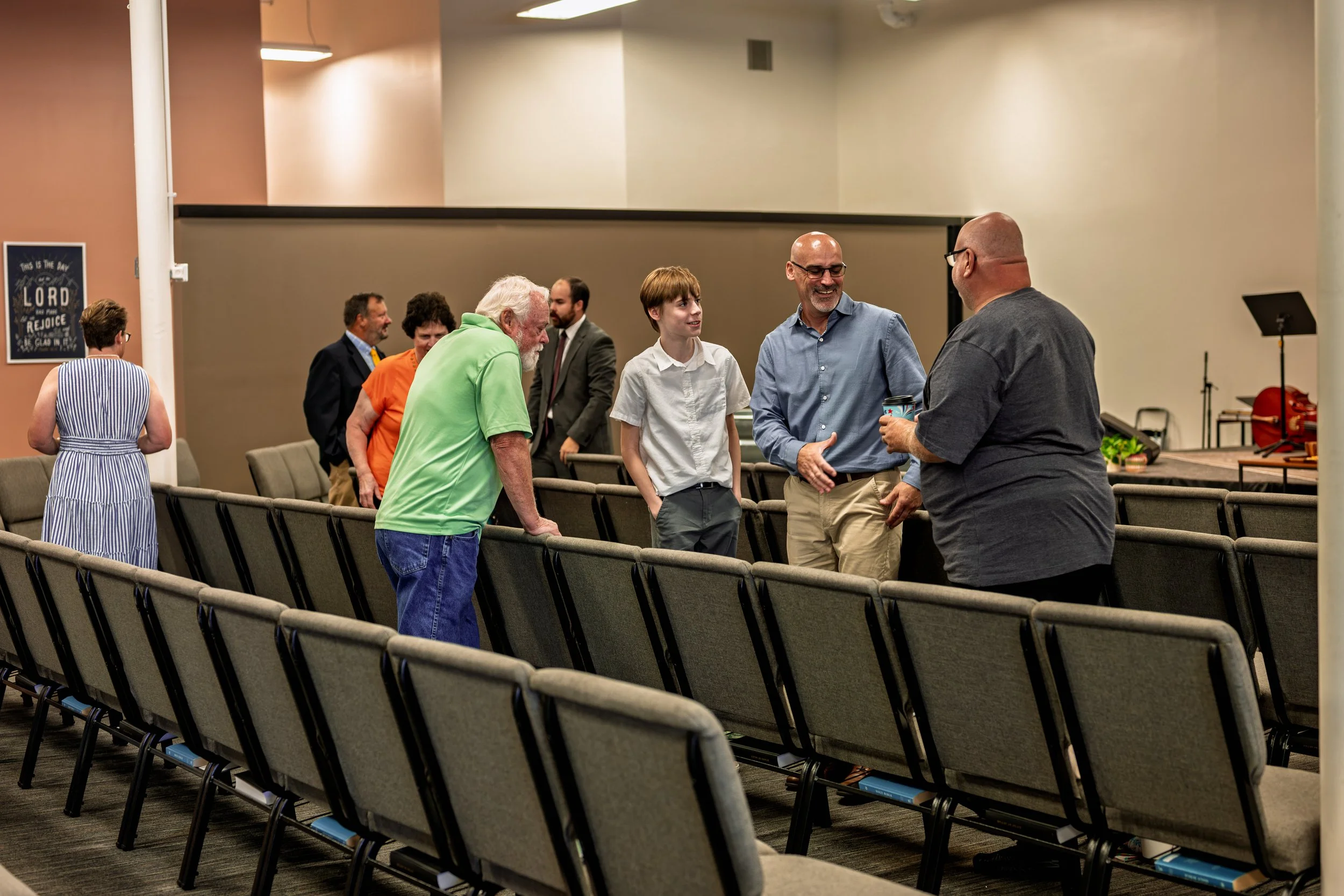 Group of people in a church or community center, standing and talking among empty rows of chairs, with a sign on the wall that reads 'This is the day the Lord has made; let us rejoice and be glad in it.'
