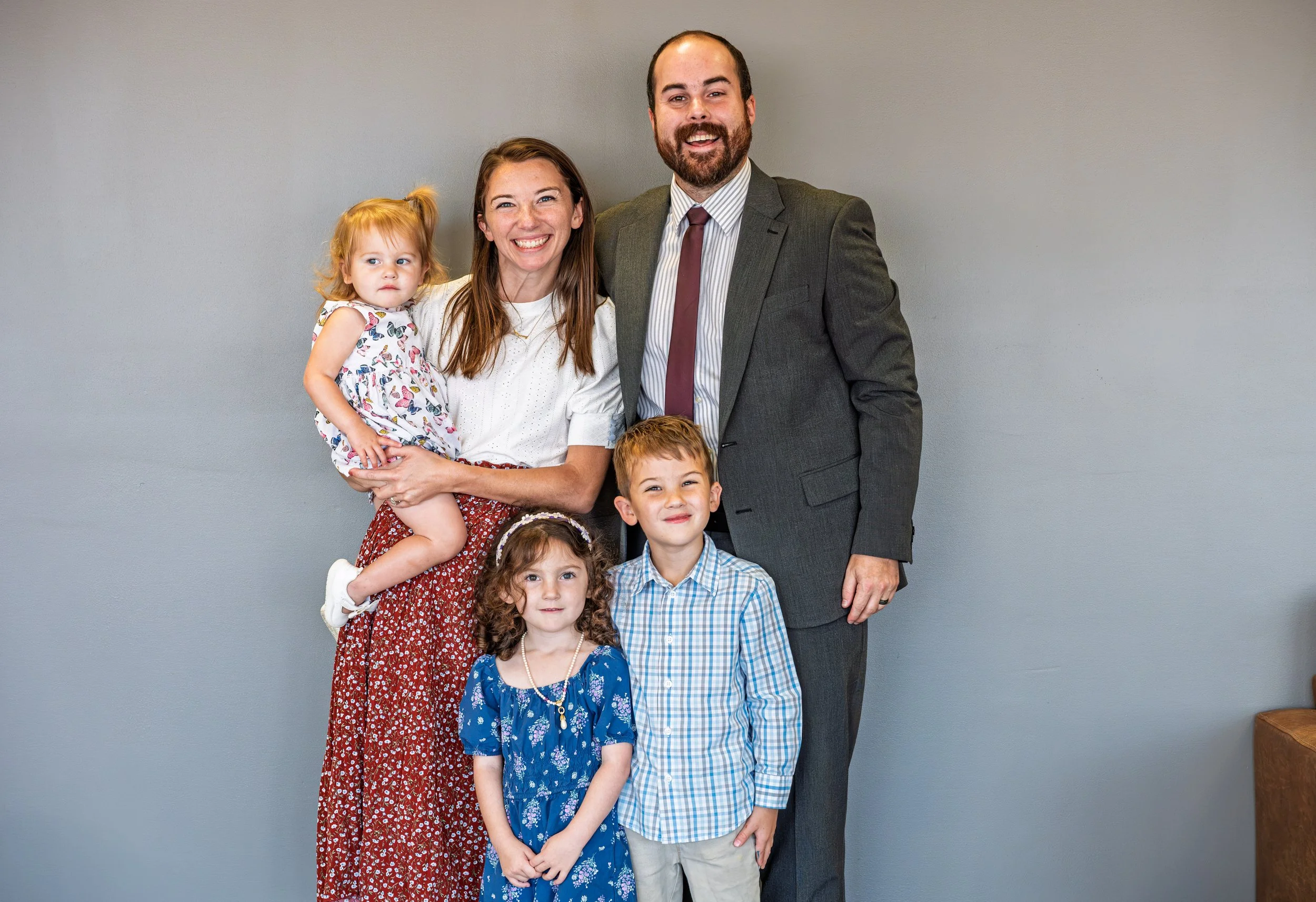 A family of six people, including a man in a suit, a woman, and four children, standing together against a plain gray wall, smiling and posing for the photo.