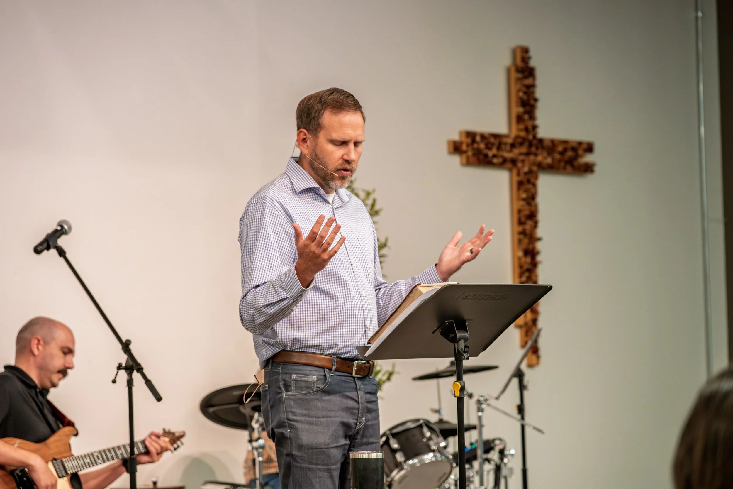 A man standing at a music stand with his eyes closed, hands raised, across from musicians playing guitars and drums, in a church with a cross on the wall.
