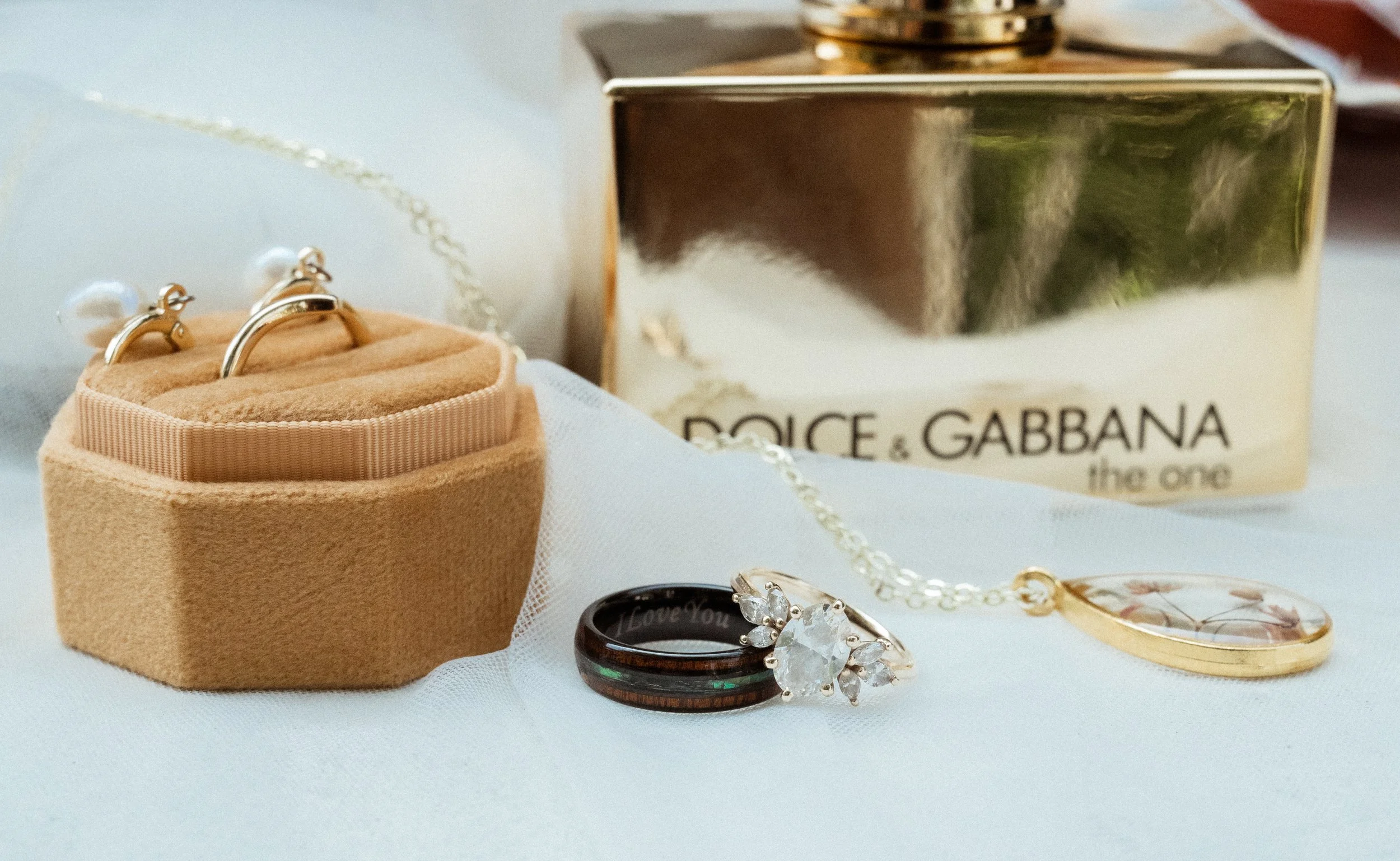 Close-up of jewelry including rings, necklaces, and earrings on a white surface with a Dolce & Gabbana perfume box in the background.