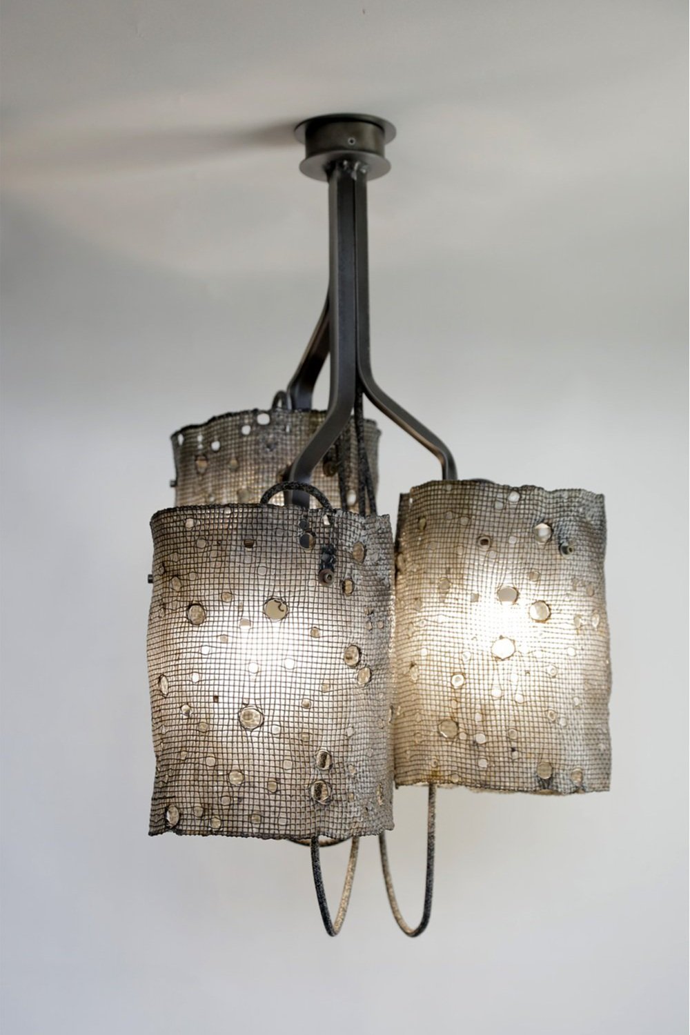Chandelier with steel frame and curved branches supporting three lit cylindrical mesh shades. Circular holes cast delicate patterns on walls and ceiling, highlighting its industrial-inspired design.