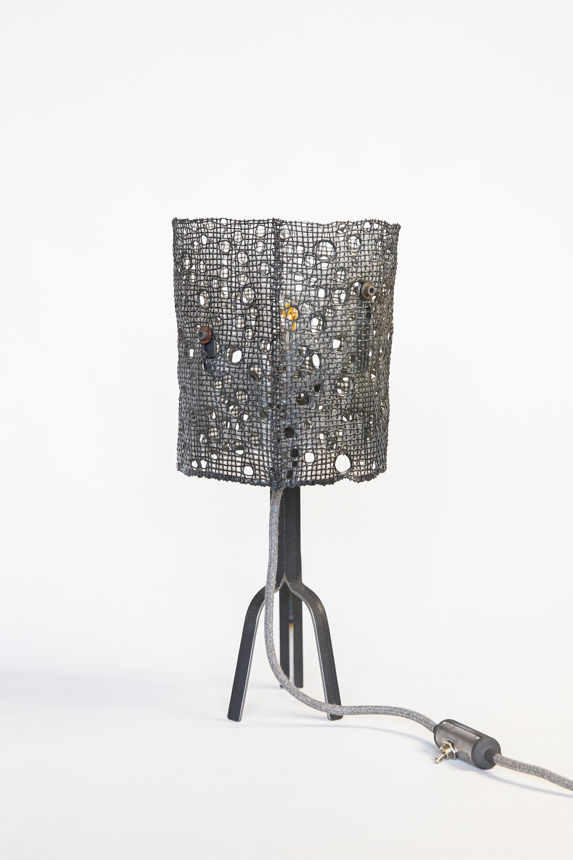 Stylish lamp with black cylindrical mesh shade featuring circular hole patterns on a sleek tripod stand with three thin legs. Fabric grey power cord with inline switch, set against a white backdrop.