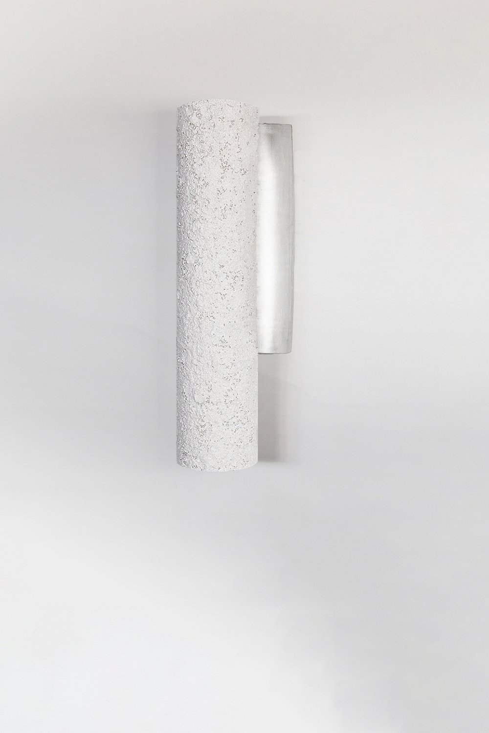 Cylindrical wall light with a rough, stone texture mounted on a plain white wall. The fixture's minimalist design contrasts its textured surface with a smooth, metallic backplate.
