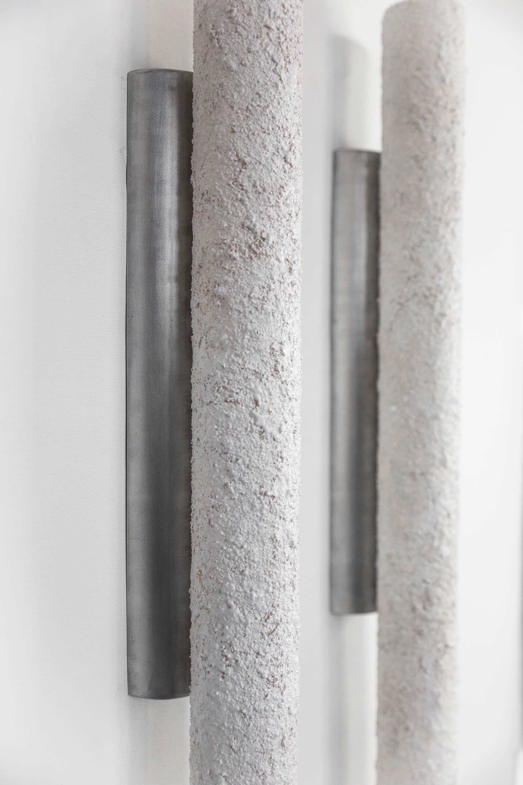 Two vertical cylindrical tube sconces with stone-like textured surfaces on a light-coloured wall. Unlit, they emphasize minimalist design and industrial style against a neutral background.
