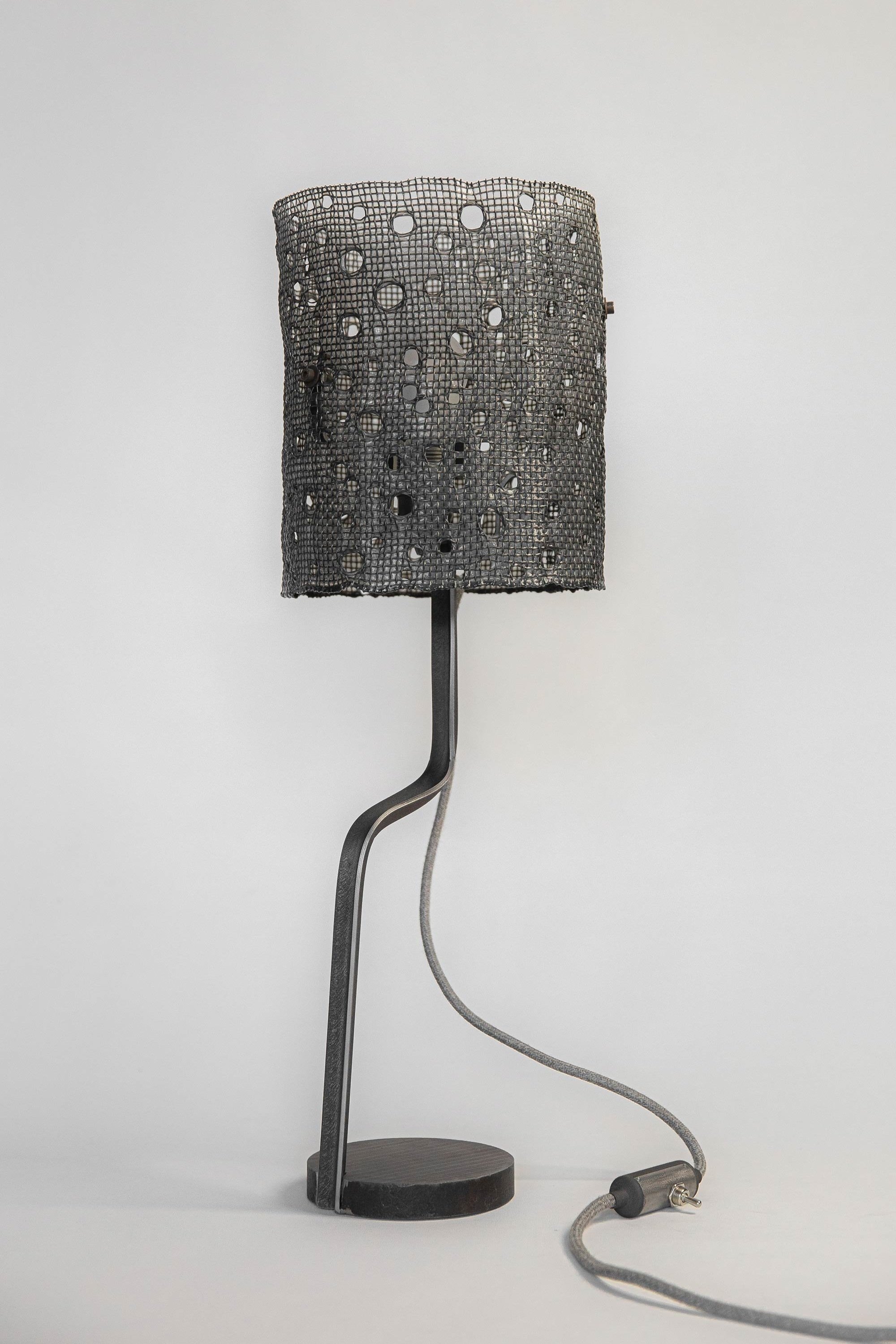 Modern table lamp with an unlit cylindrical mesh shade featuring circular hole patterns. Supported by a curved steel stand and round base, it has a fabric-covered grey cord and minimalist aesthetic.