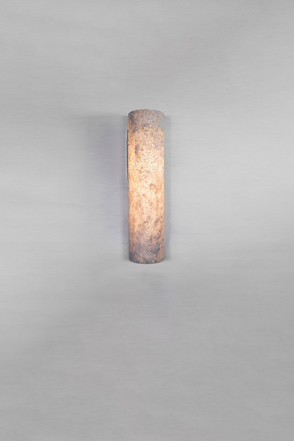 Wall-mounted cylindrical light fixture with a textured stone-like surface emitting a warm, diffused glow. Set against a plain background, the design showcases rugged yet elegant craftsmanship.