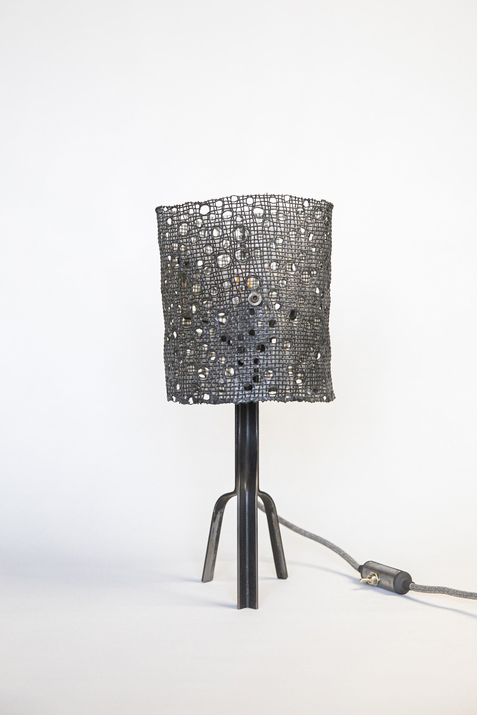 Unique lamp with black mesh cylindrical shade featuring circular holes, sleek steel tripod stand. Includes a grey cylindrical switch on the textile power cord, highlighted against a white backdrop.