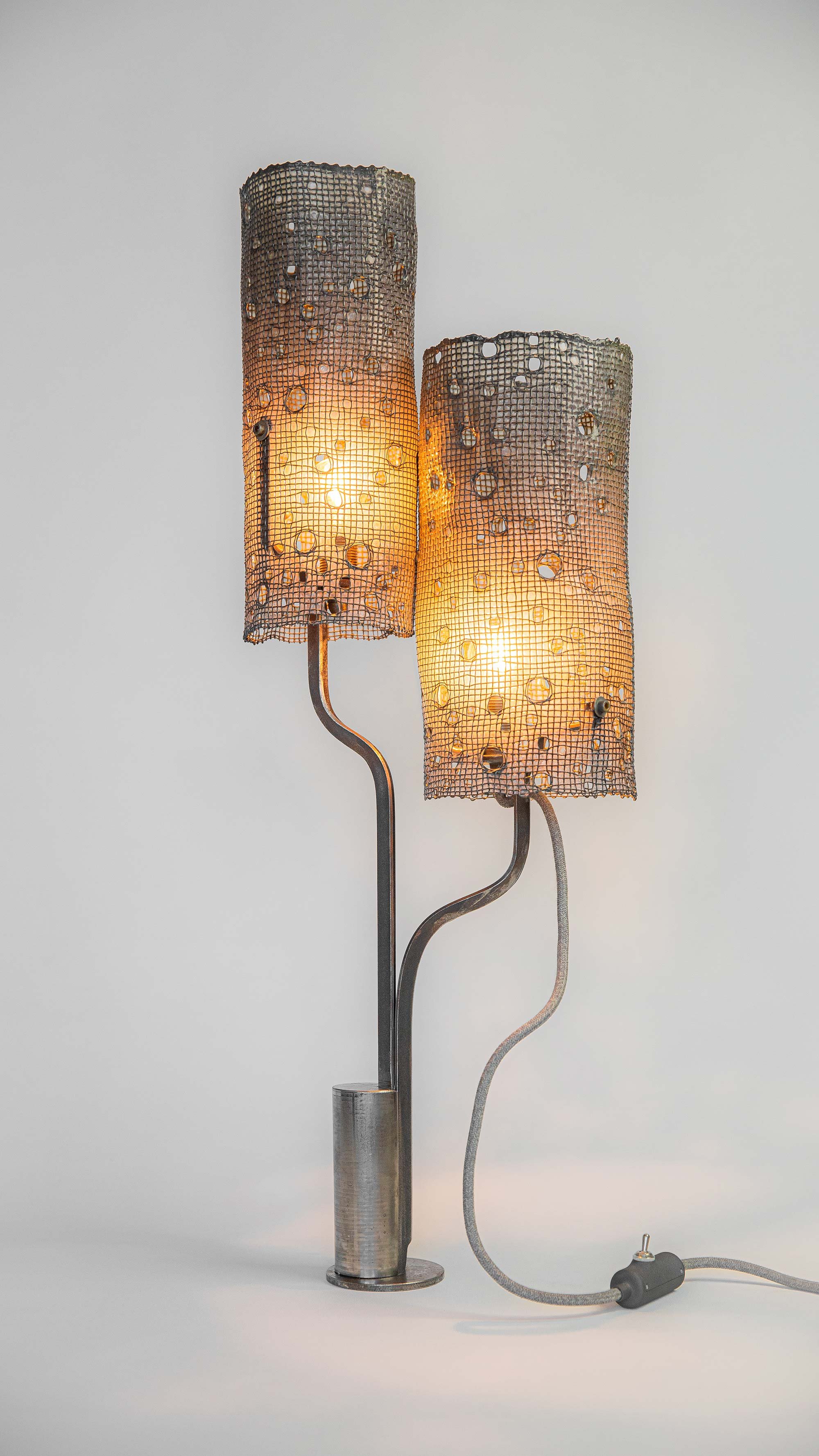 Modern lamp with two cylindrical mesh steel shades featuring small holes. Curved metal stems connect to a cylindrical base. Textile cable with inline switch blends industrial and contemporary styles.
