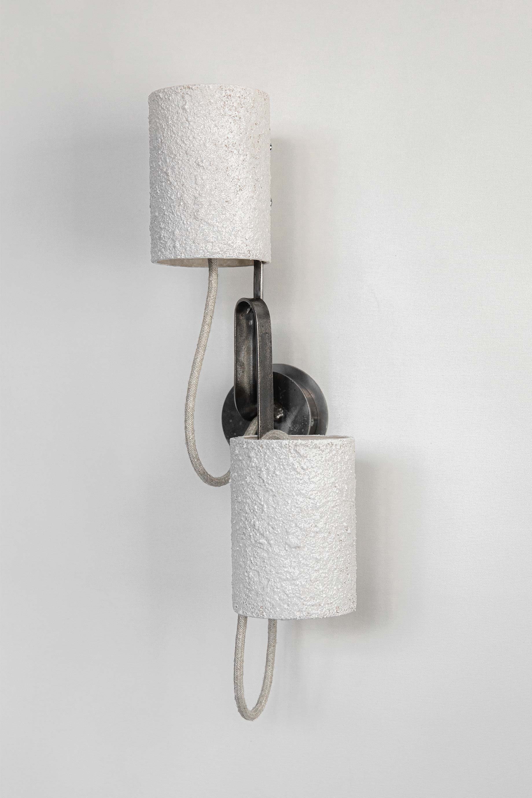Wall-mounted light with two textured white cylindrical shades on curved metallic arms. A looped cord adds industrial charm, while its modern aesthetic shines as an unlit decorative piece.