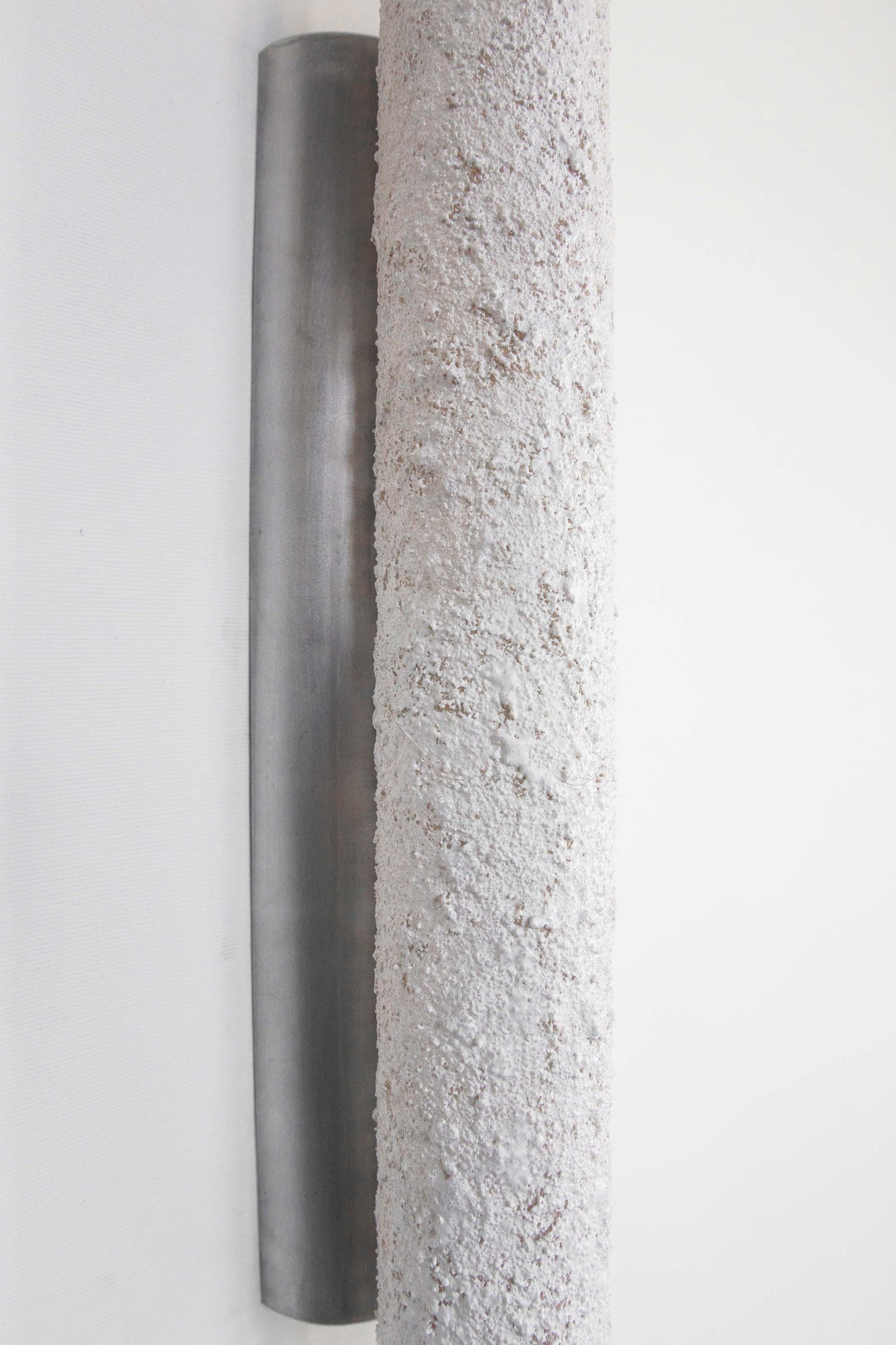 Close-up of a wall-mounted tube light fixture with an unlit textured stone-like surface. Intricate detailing contrasts with the sleek brushed steel rose, blending ruggedness with refined design.