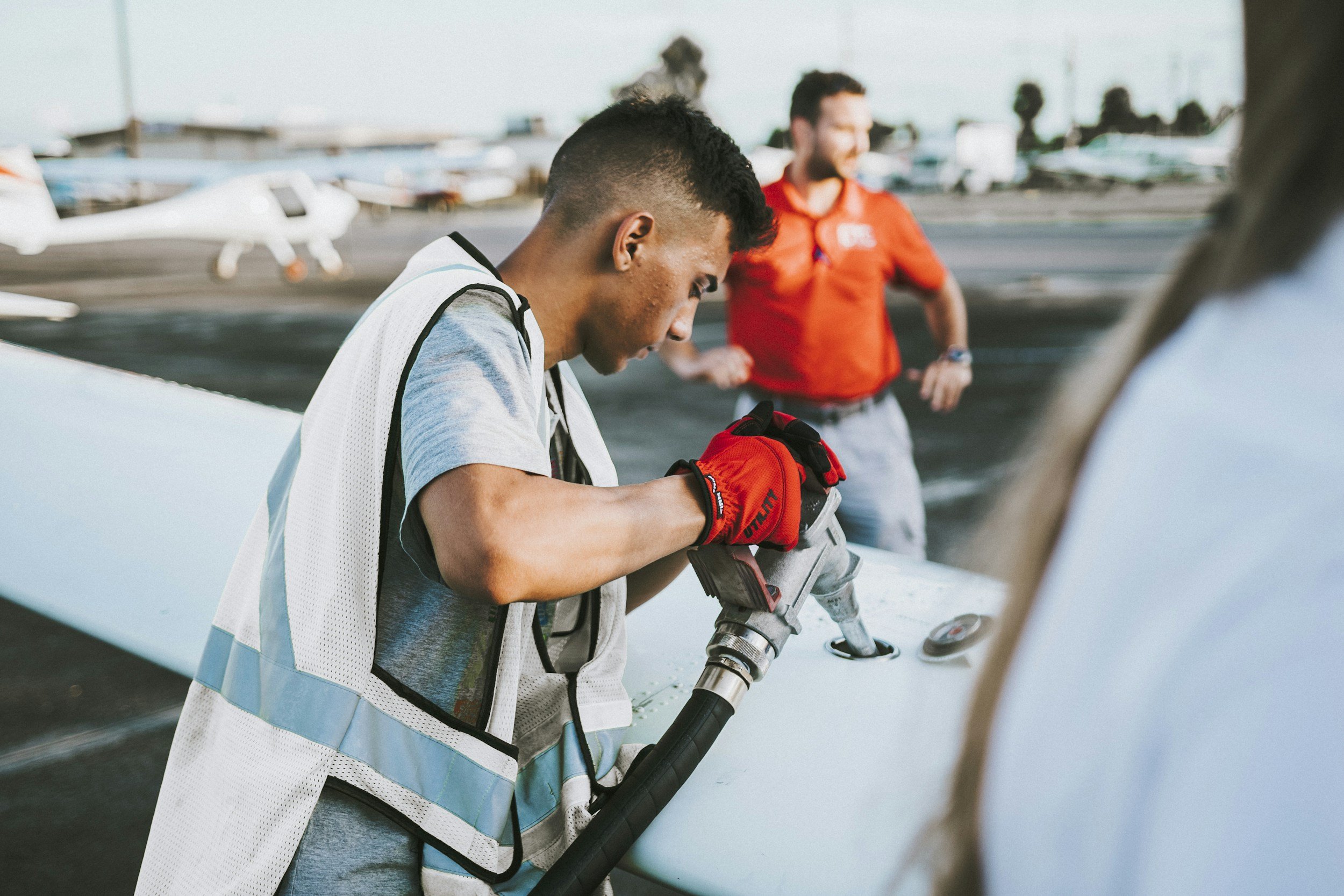 A man in a white vest with reflective stripes and red gloves working on an airplane propeller with a power drill at an airport. Another man in a red shirt stands in the background with his hands on his hips.