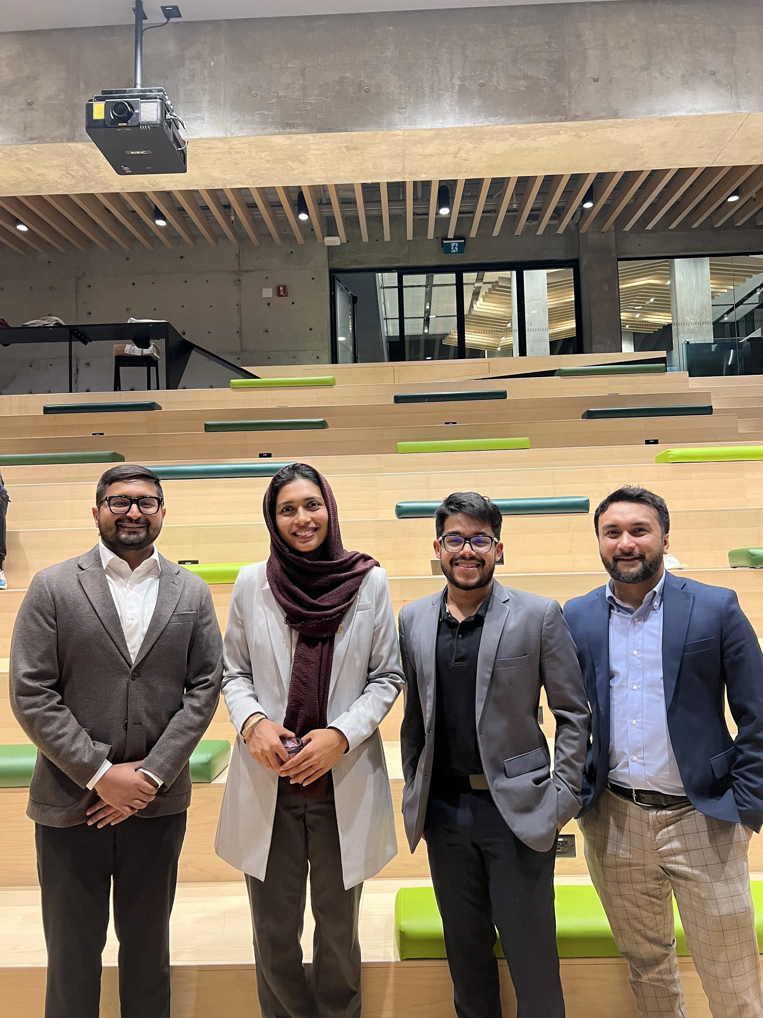 Four professionally dressed individuals smiling and standing in a modern auditorium with tiered wooden seating and green and black accents.