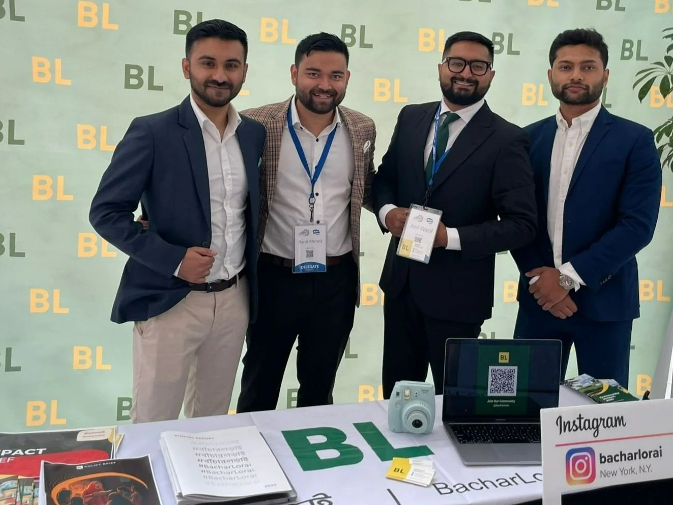 Four men in formal attire standing together at a conference booth, smiling, with a backdrop displaying the letters 'BL'. The booth has various materials like brochures, a laptop, a camera, and signage including an Instagram handle, at an event in New York.