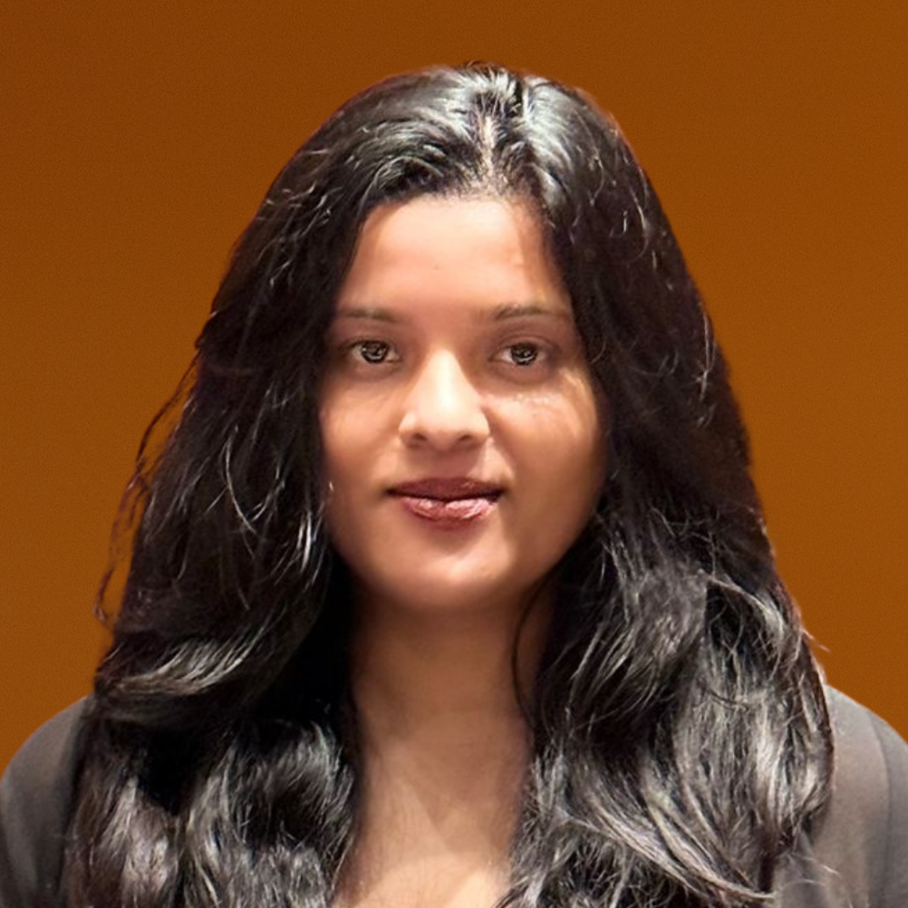 A young woman with long dark hair smiling in front of a blurred orange background, wearing a gray winter jacket with a fur-lined hood.