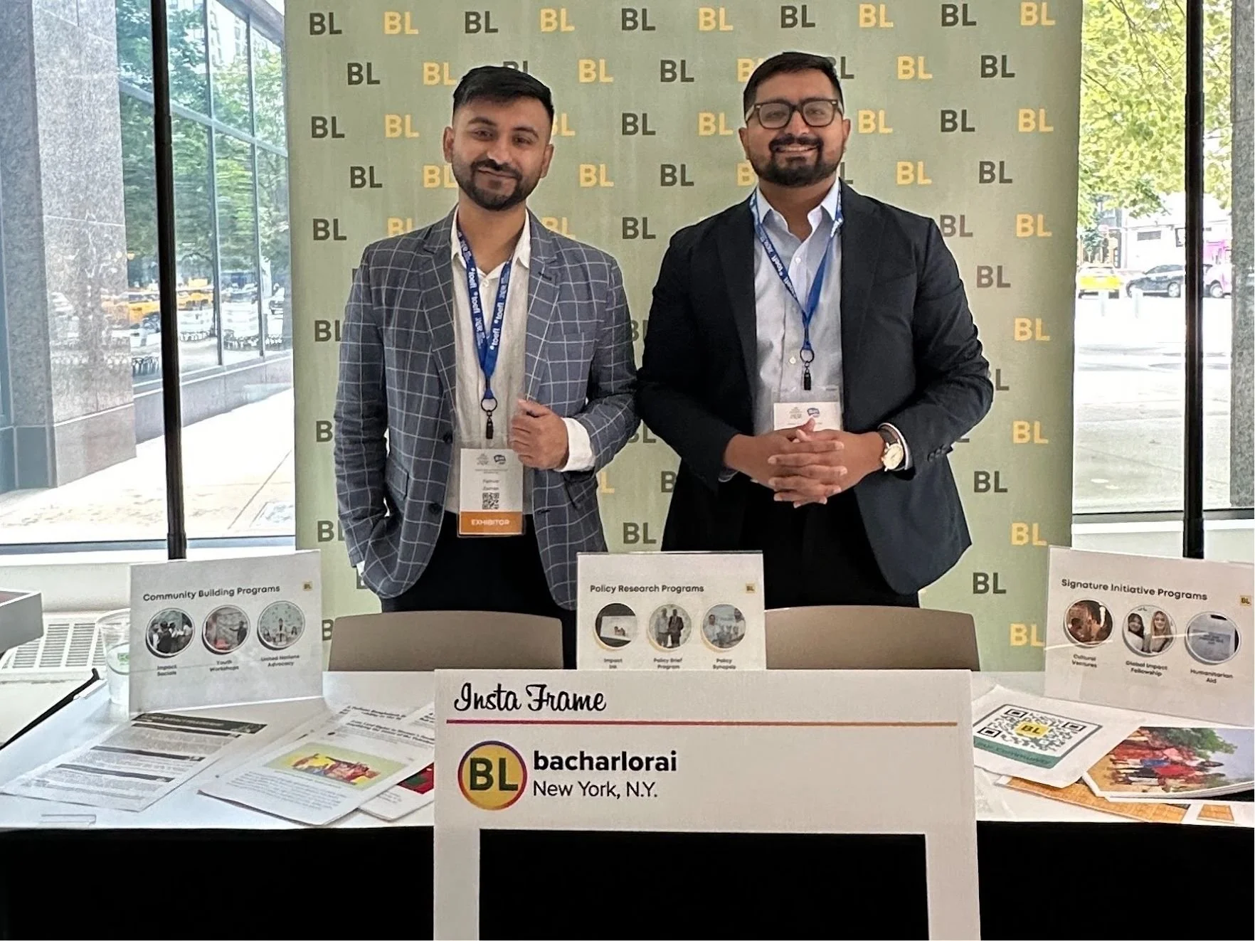 Two men in business attire standing behind a table at a conference, smiling at the camera. The table displays brochures and promotional materials for Bacharach, New York, N.Y., with a backdrop featuring the BL logo repeatedly.