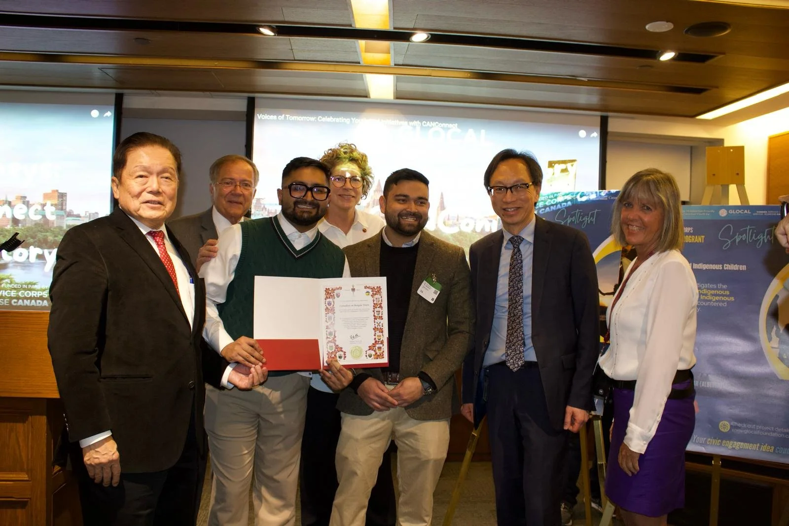 Group of eight diverse people standing together in a room, holding a certificate, with presentation screens and banners in the background, celebrating an achievement.