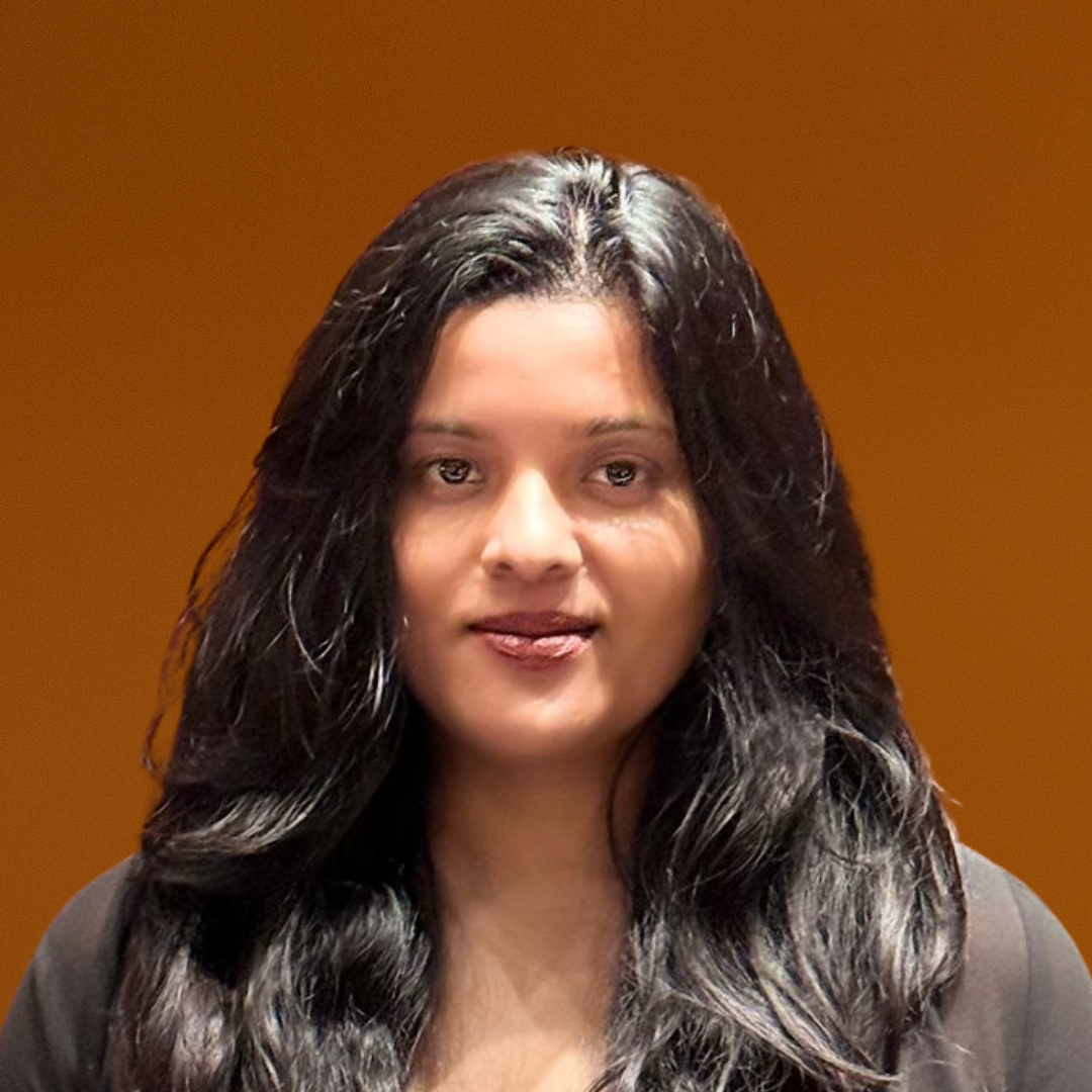 A young woman with long dark hair smiling in front of a blurred orange background, wearing a gray winter jacket with a fur-lined hood.